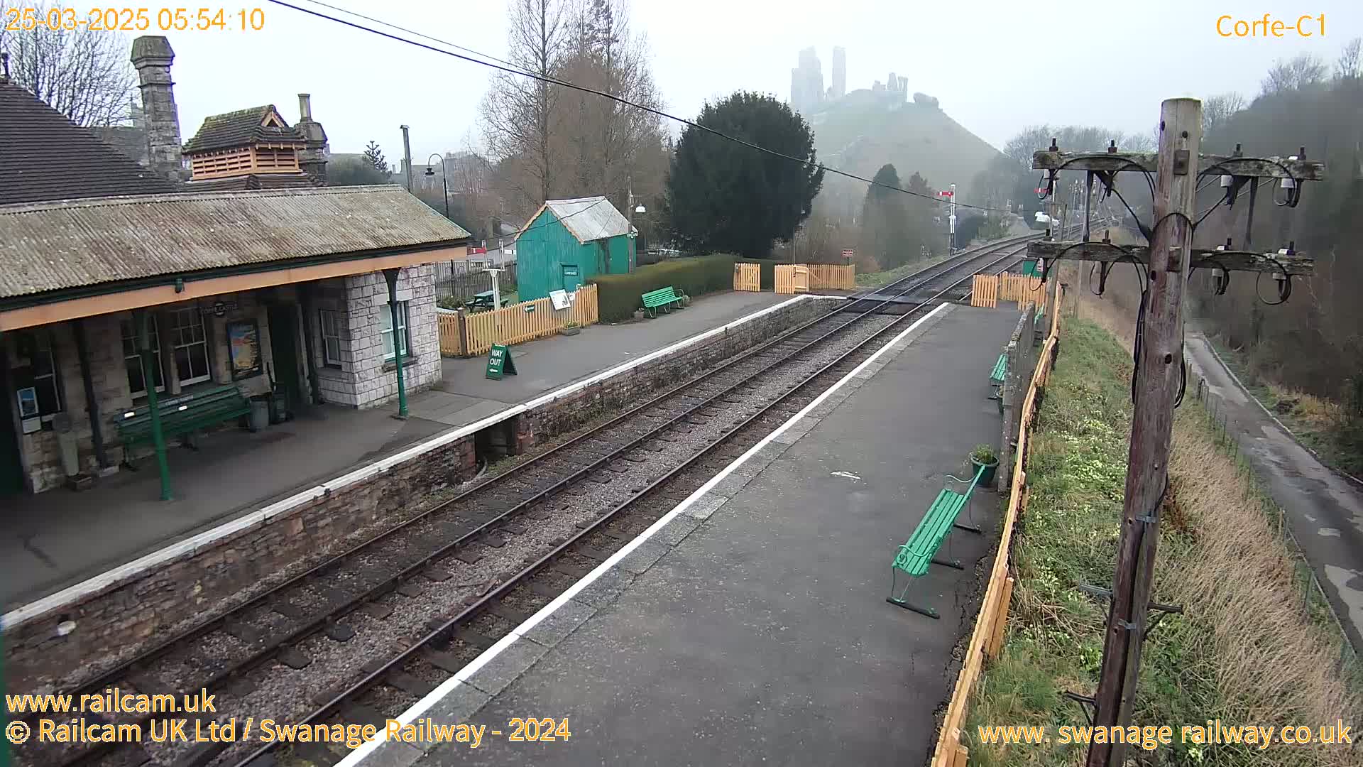 A train station platform with a stone building, benches, and railway tracks, overlooks a hill with a castle in the misty distance.