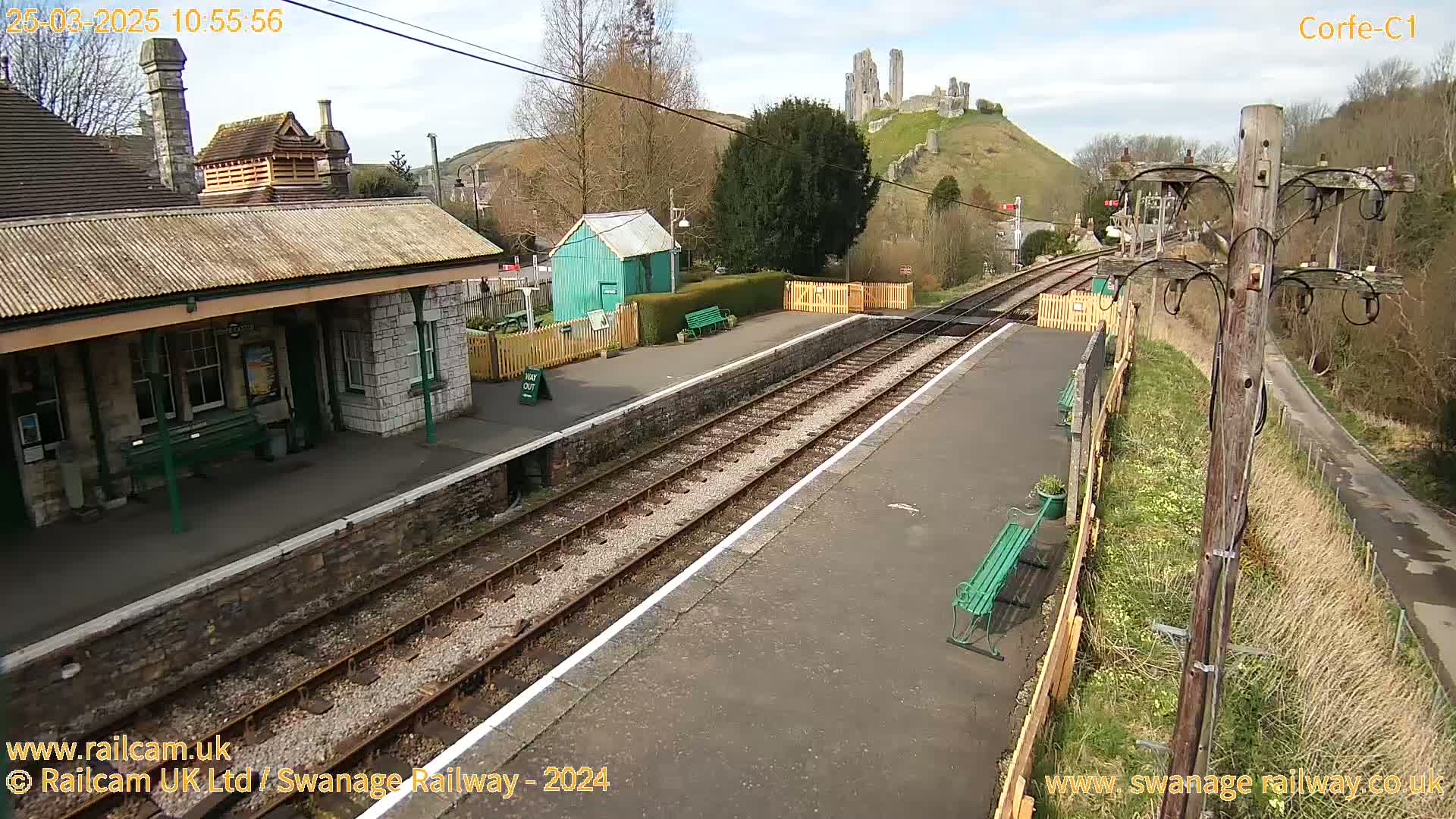 A train station with a stone building, a small green shed, and train tracks, all under a partly cloudy sky, with a hilltop castle in the background.