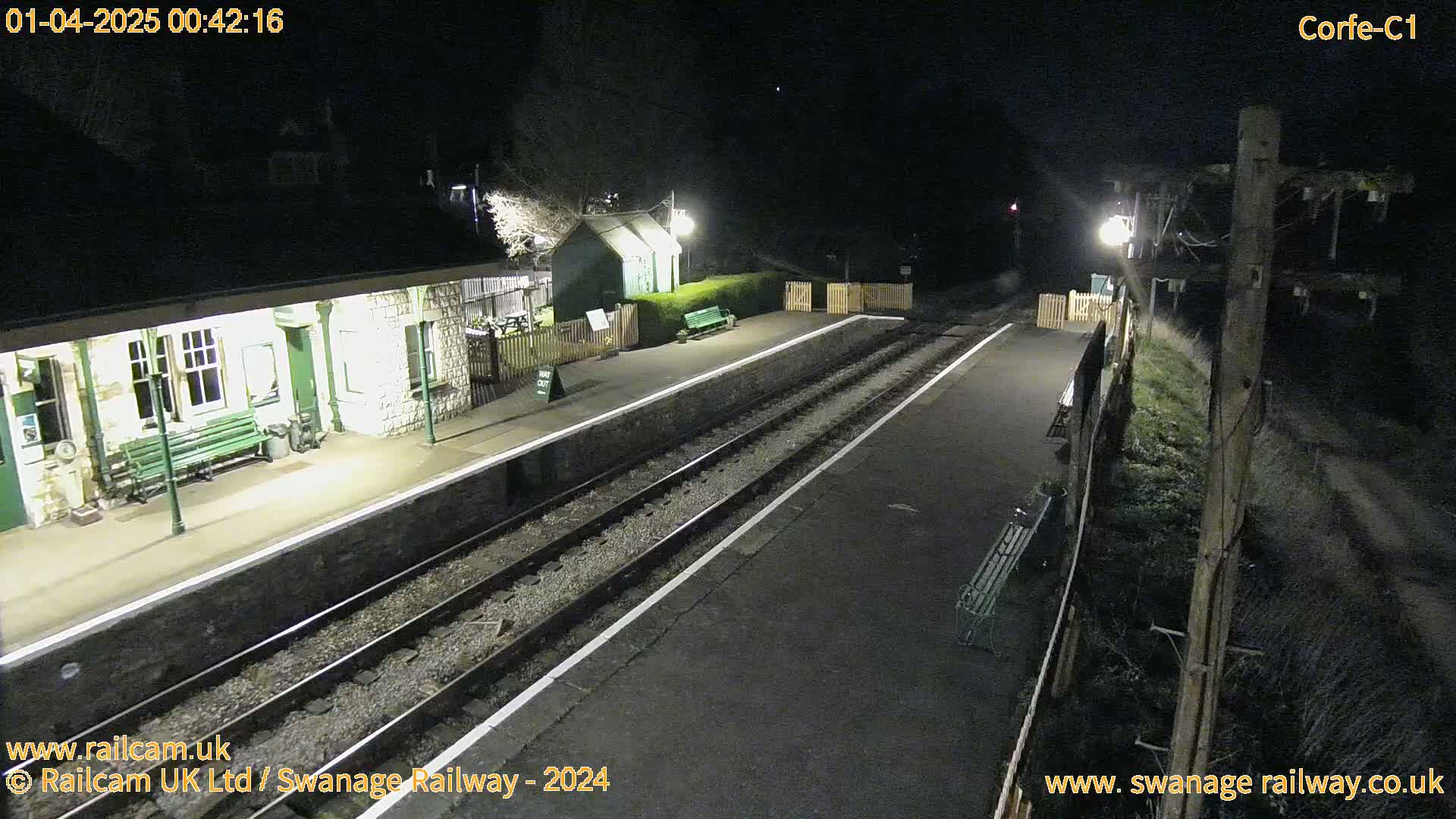 A nighttime view of a deserted train station platform with two sets of train tracks, a small building, and benches under dim lighting.