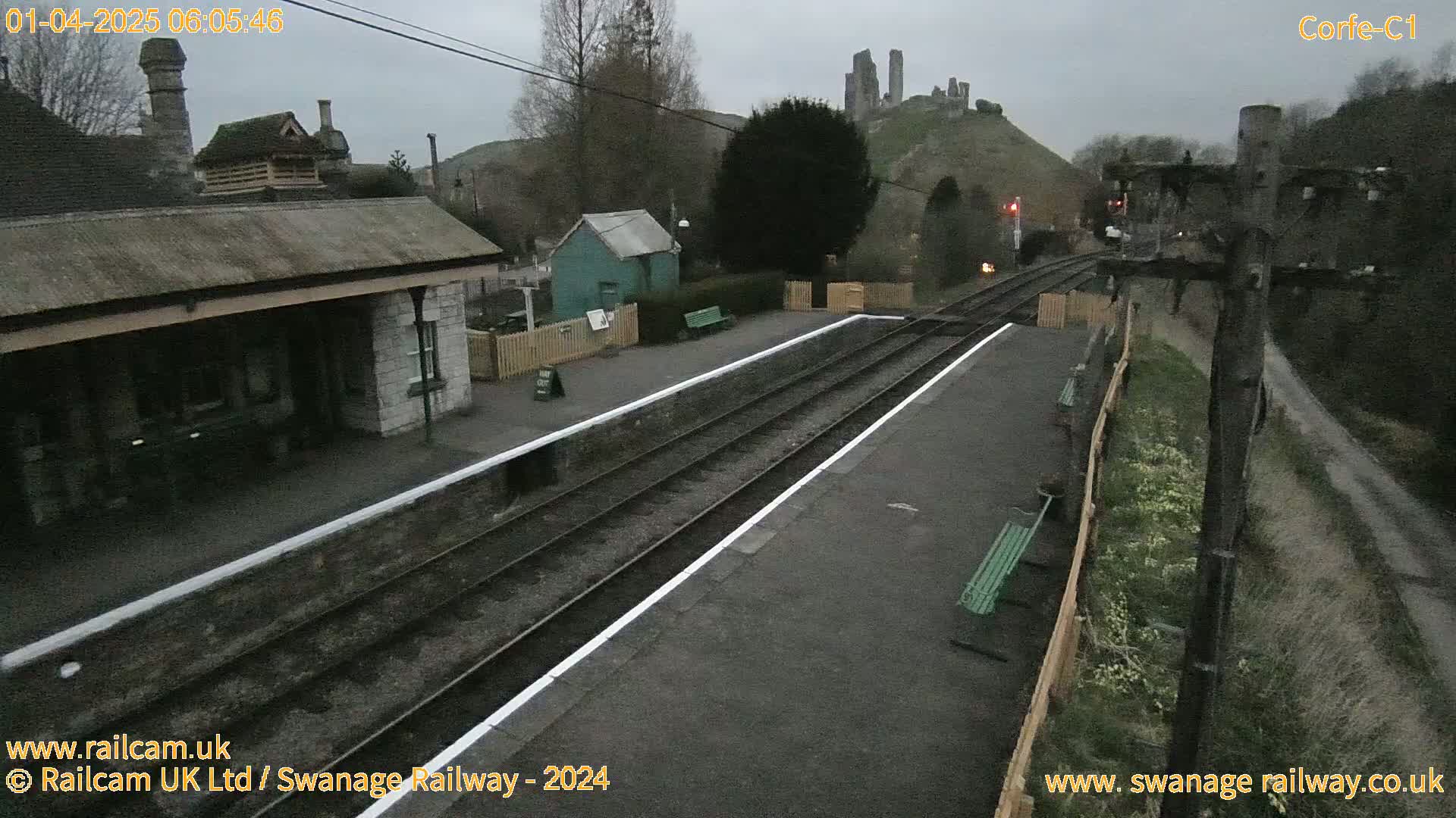 A train station platform with two sets of tracks under an overcast sky, featuring a stone building and a distant castle ruin on a hill.