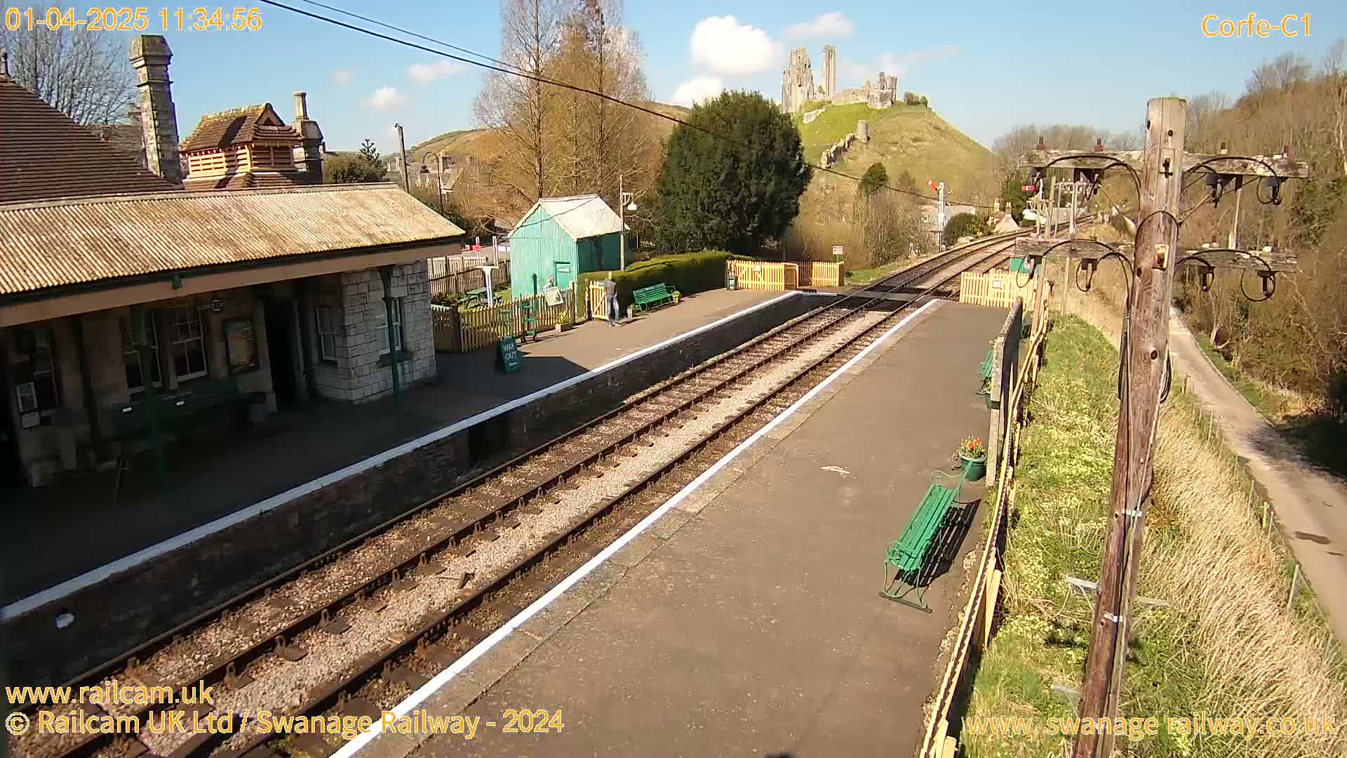 A train station platform with two tracks, a stone building, and a hilltop castle in the background under a sunny sky.