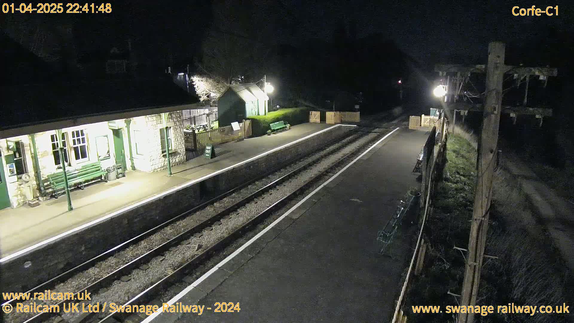 A nighttime view of a deserted train station platform with two train tracks and a small building under artificial light.