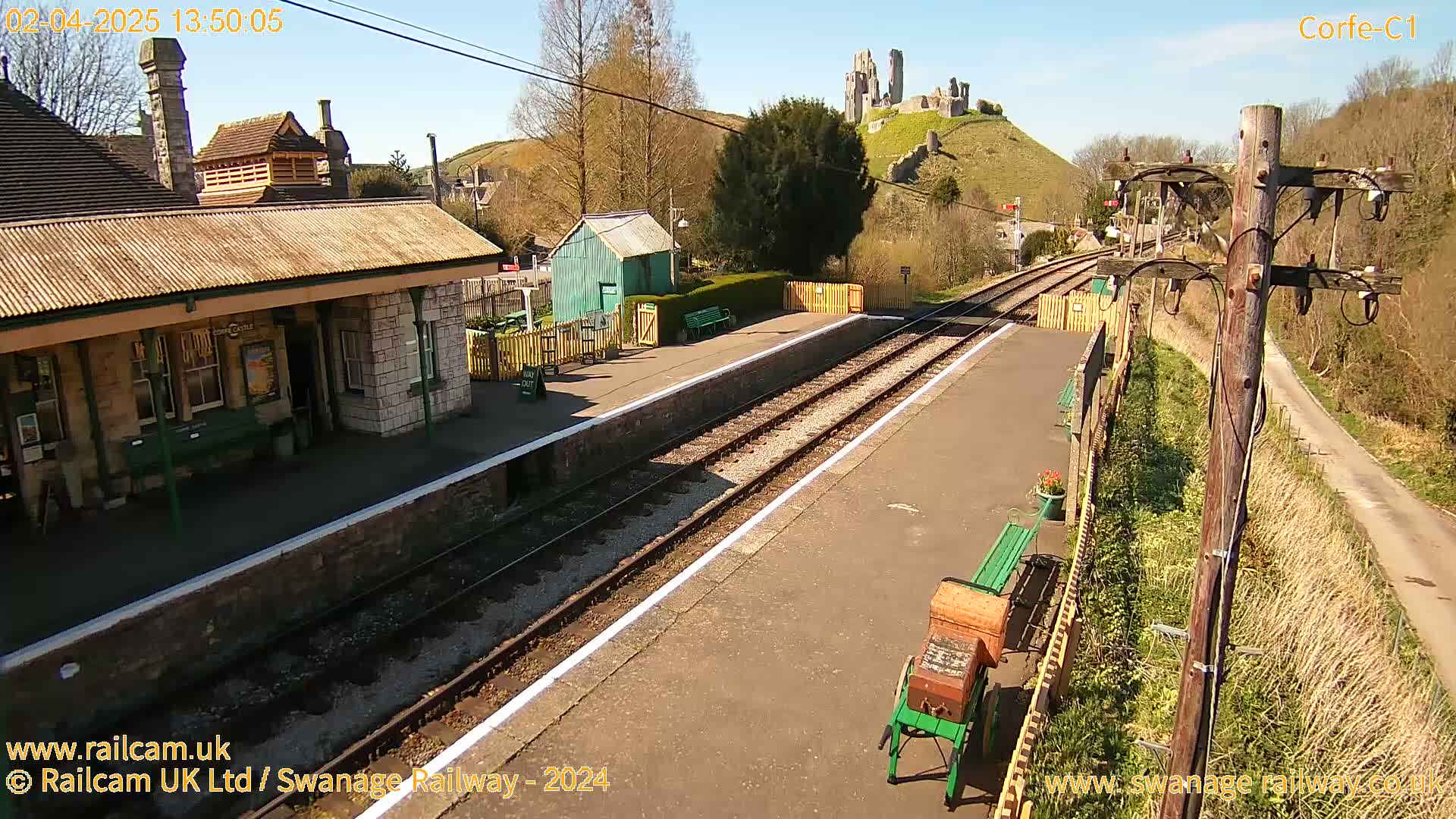 A train station platform with a stone building and a small green shed, sits beneath a sunny sky and a hill topped with castle ruins.