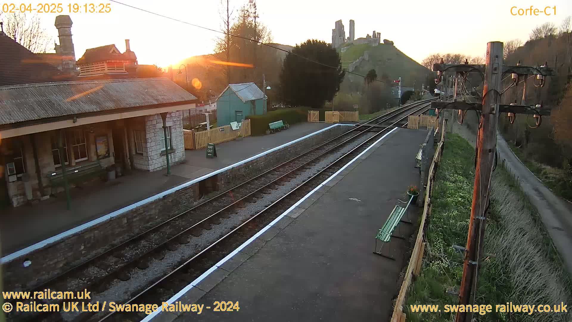 A train station with two tracks and a stone building is shown at sunset, with a hilltop castle in the background.