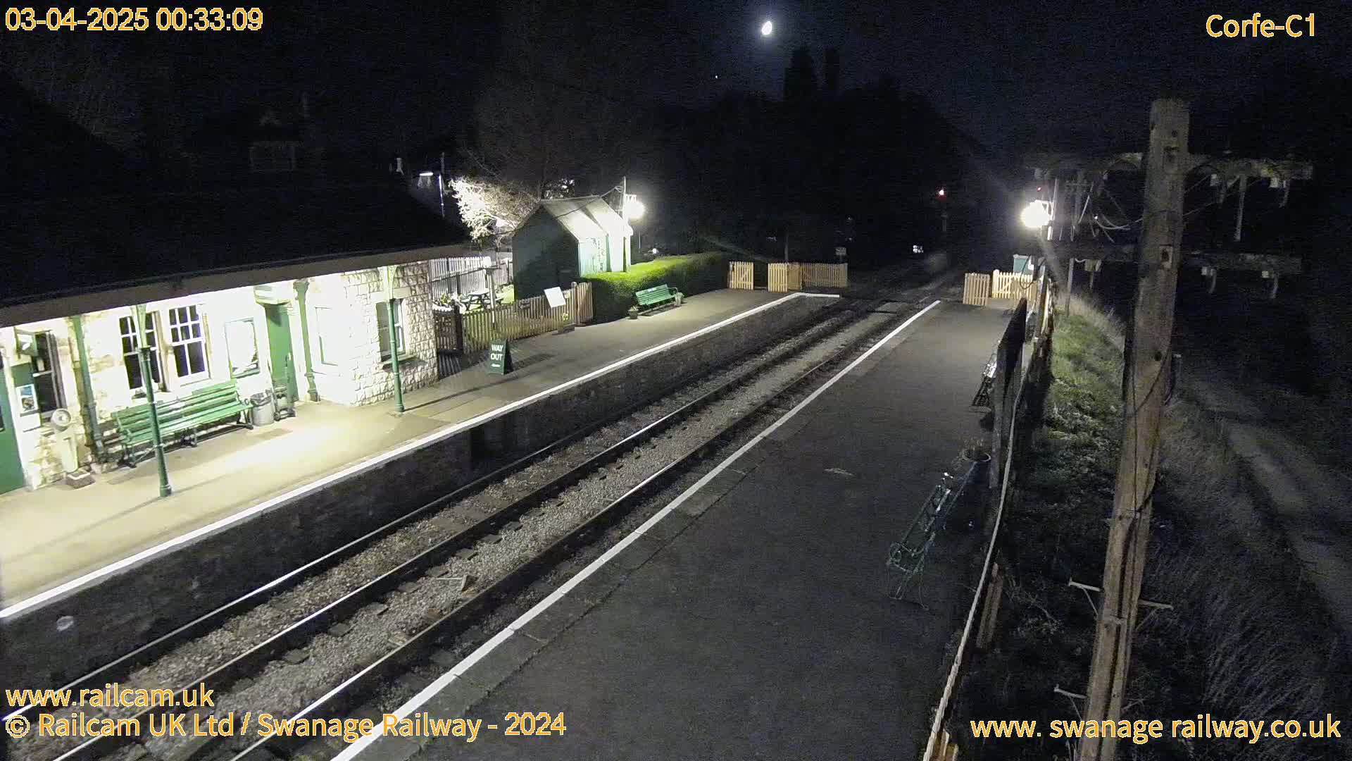 A mostly empty train station platform is illuminated at night under a bright moon.