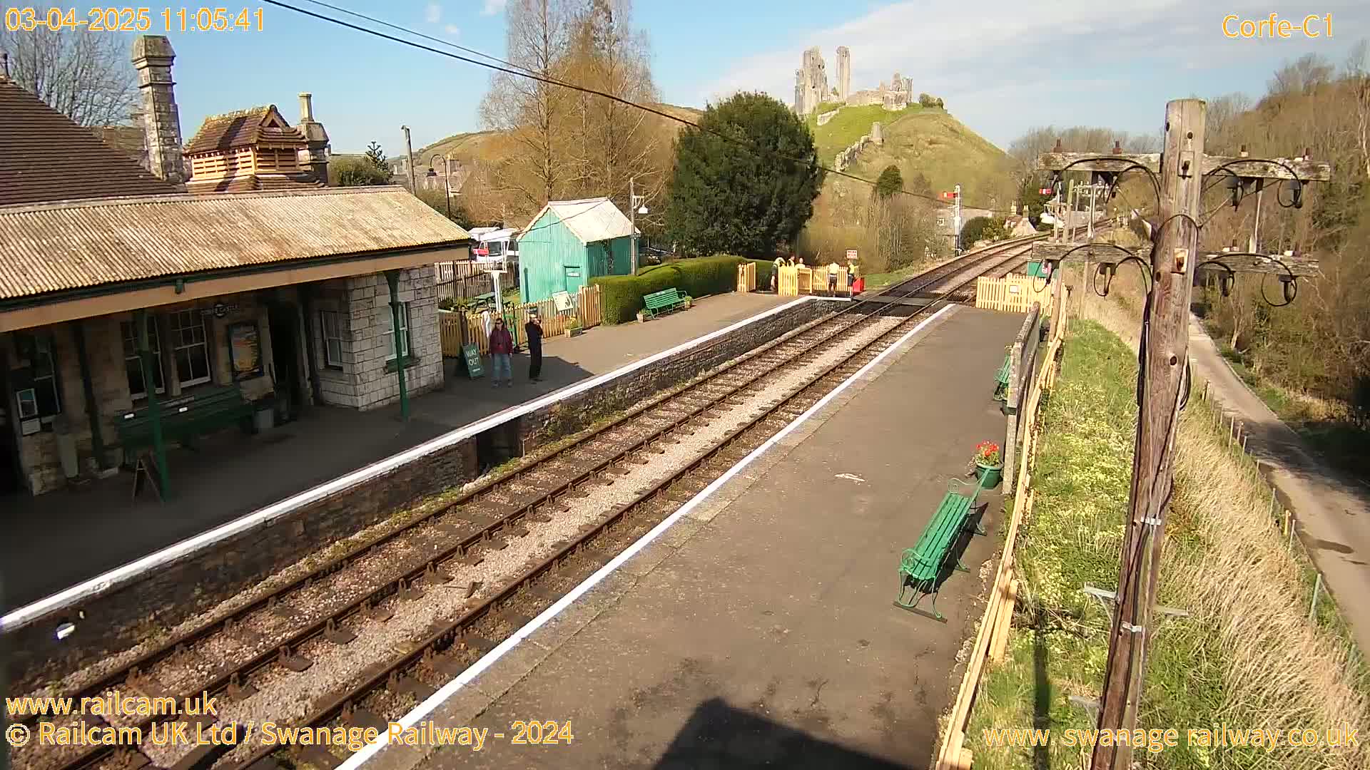 A train station platform with a stone building, a small green shed, and a hill with ruins in the background under a sunny sky.