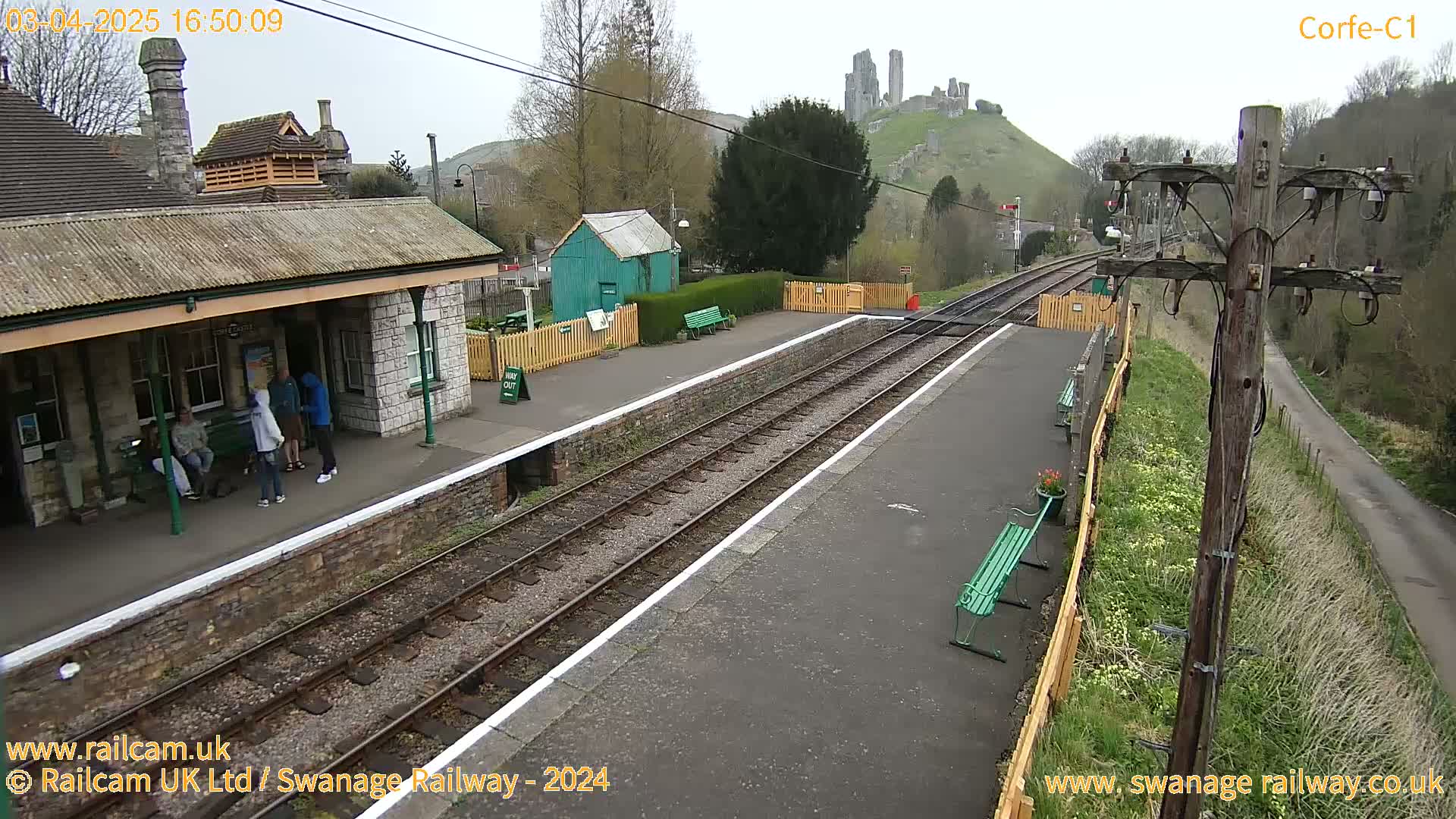 A railway station platform with several people standing near a stone building, train tracks extending into the distance, and a castle visible on a hill in the background under an overcast sky.