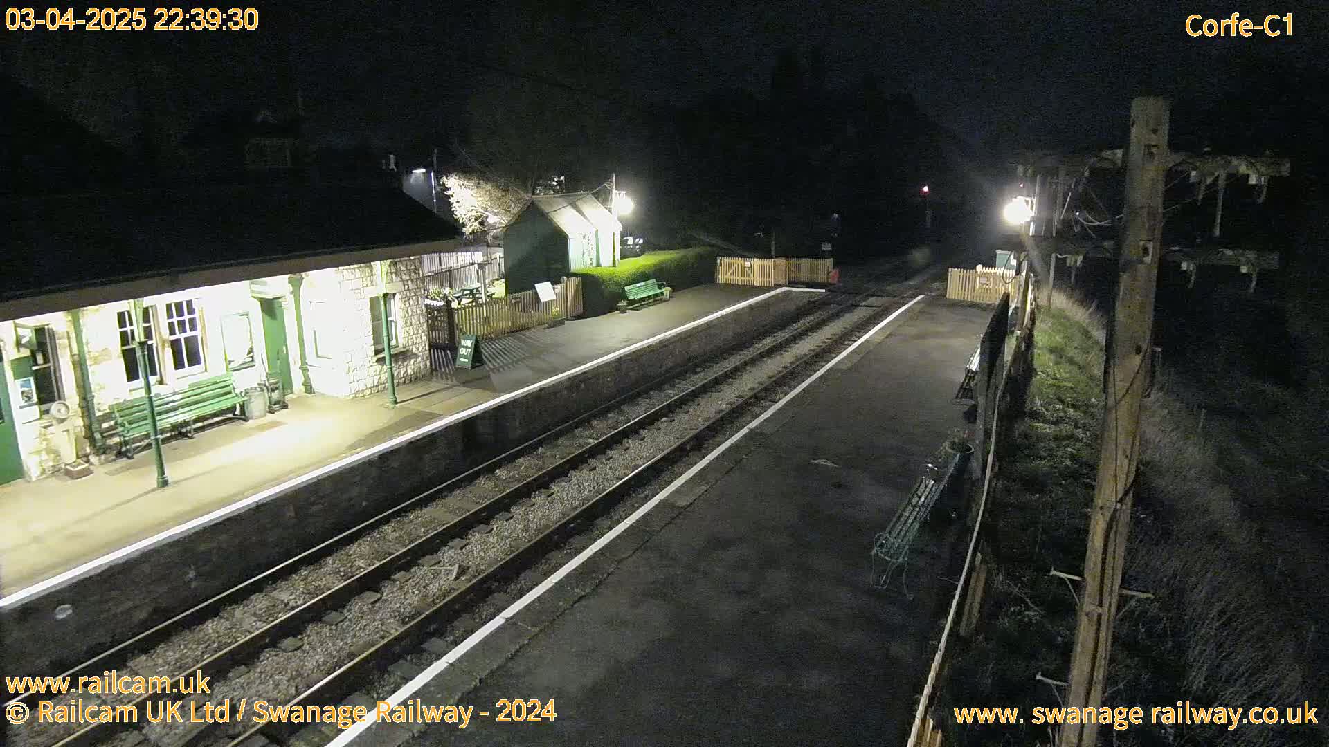 A nighttime view of a deserted train station platform with two sets of tracks and a building lit by lamps.