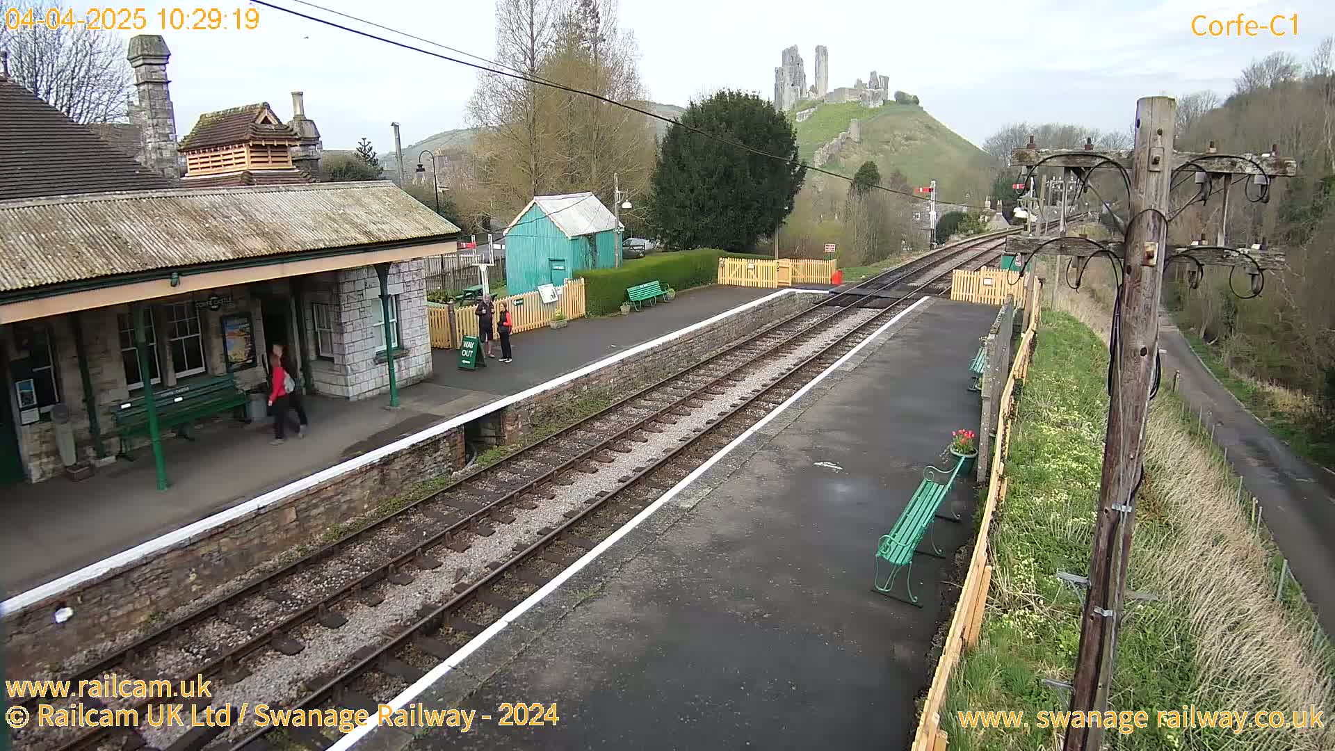 A train station platform with a stone building and a hill topped with a castle ruin in the background, under a partly cloudy sky.