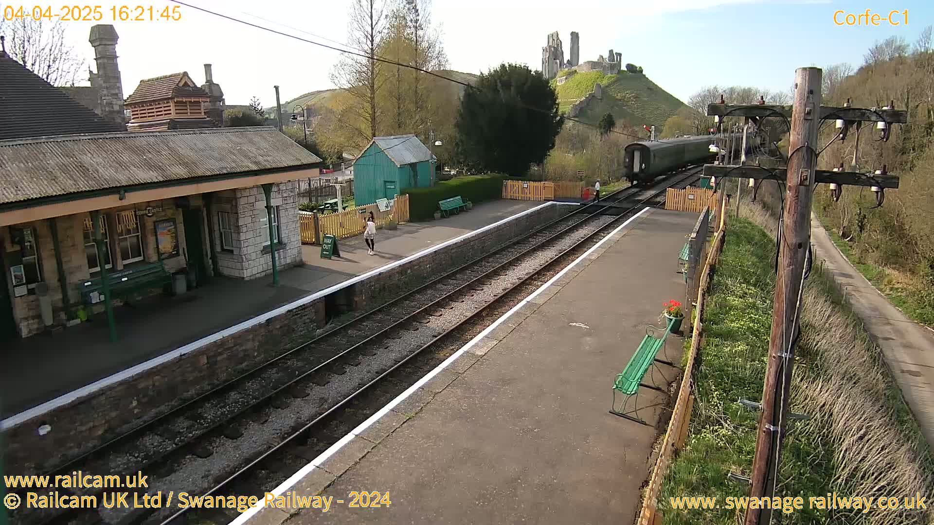 A train departs a railway station on a sunny day, with a castle visible on a hill in the background.