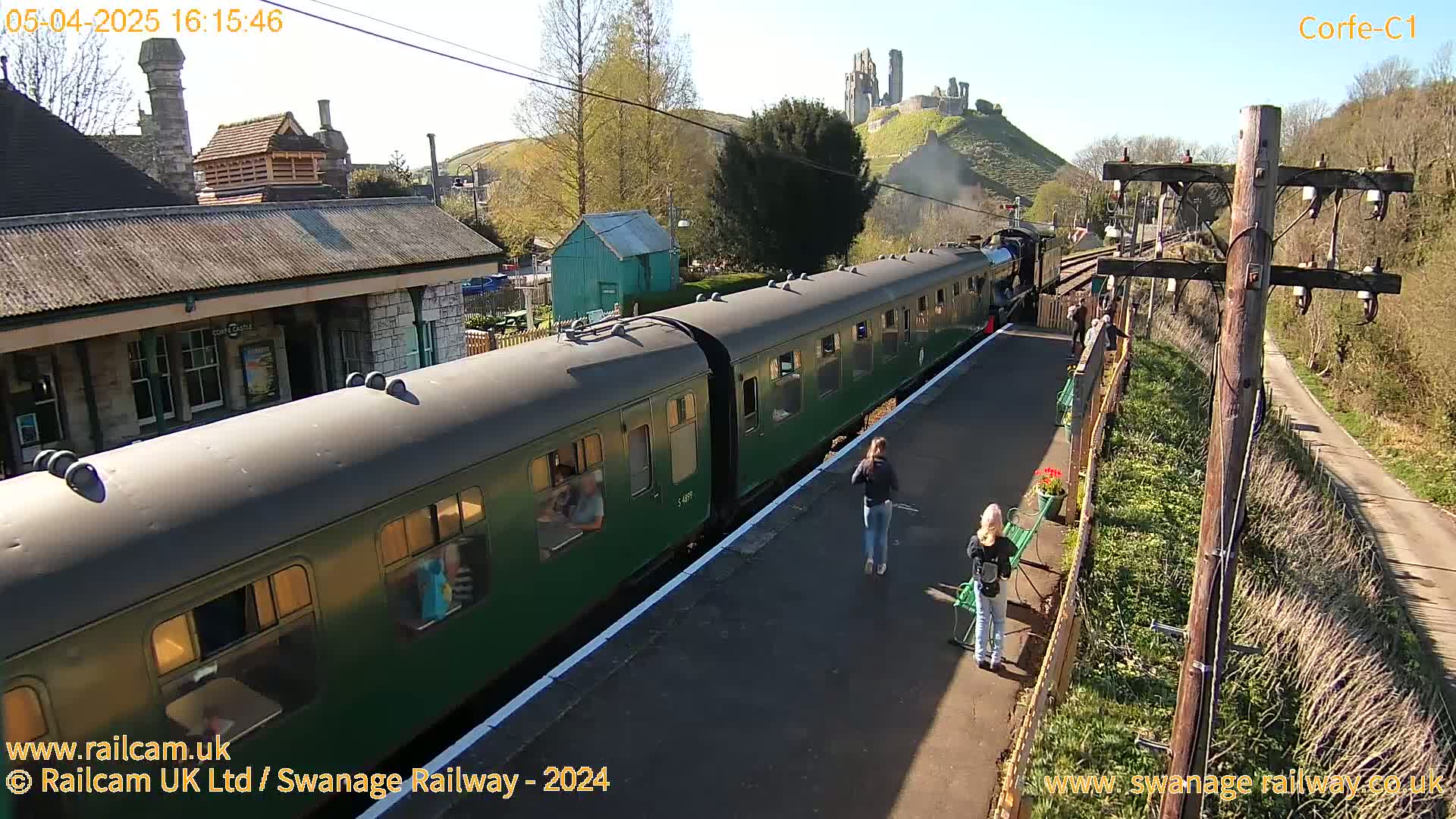 A green passenger train is at a station on a sunny day, with a hill and castle ruins visible in the background.