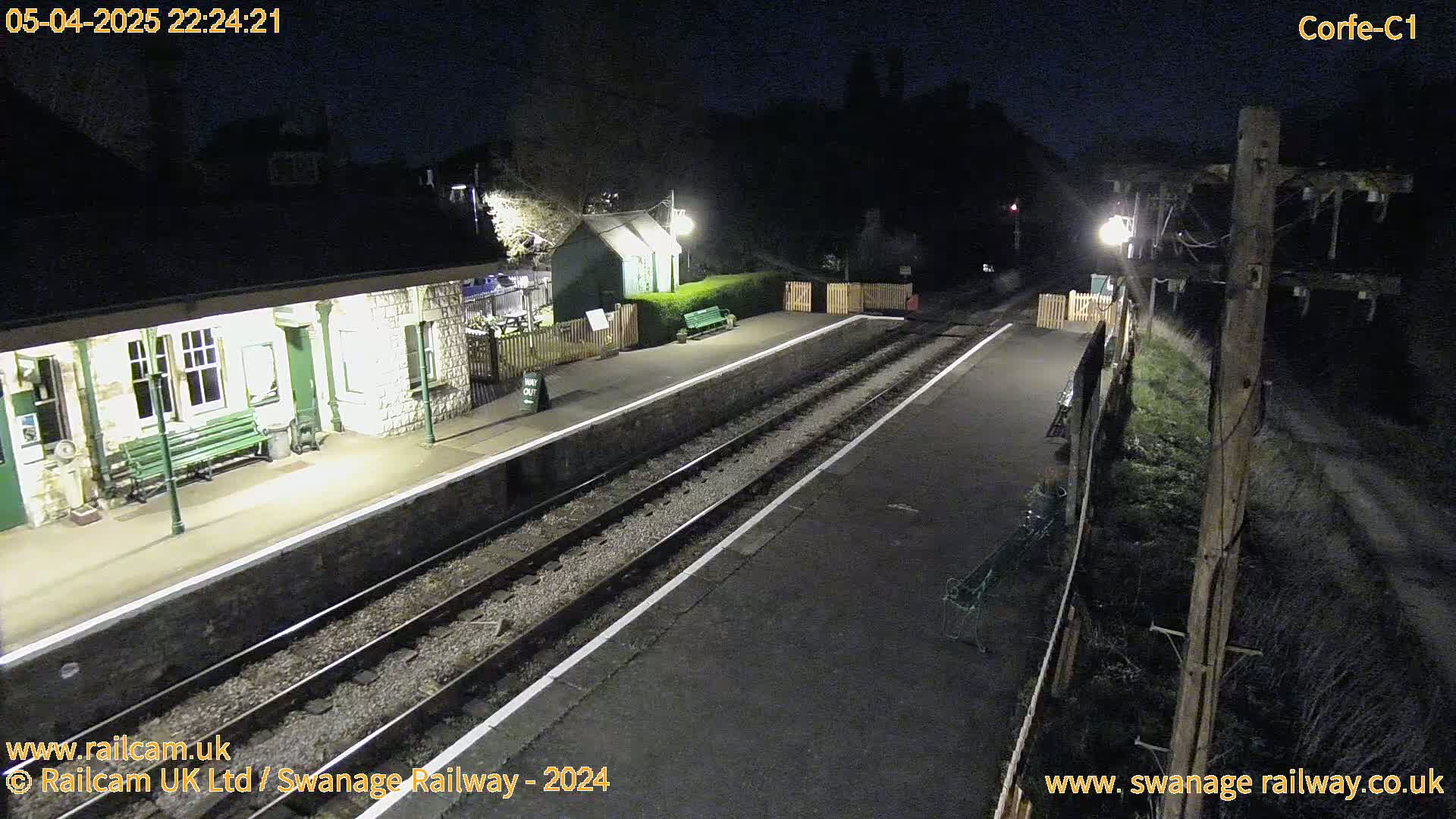 A deserted train platform at night, illuminated by artificial lights, with train tracks extending into the darkness.