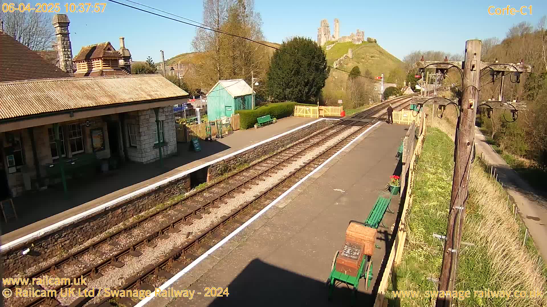 A train station platform with a stone building, a small green shed, and a distant hilltop castle under sunny skies.