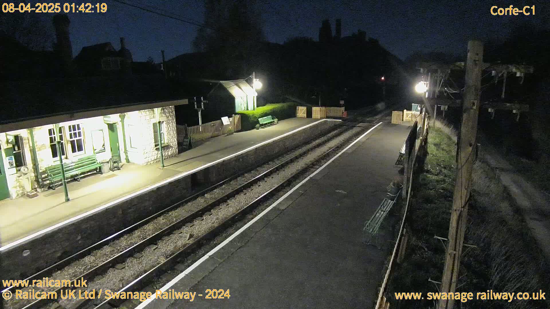 A nighttime view of a deserted train station platform with two sets of tracks, illuminated by artificial lights.