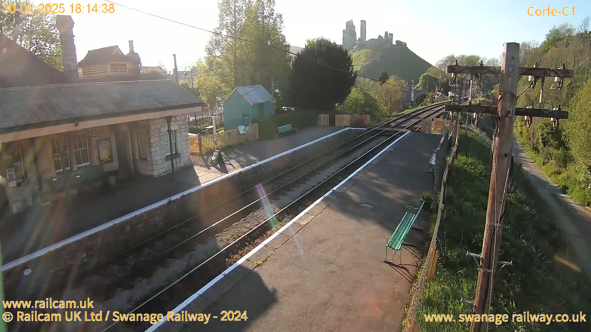 A train station platform with a stone building, a small green shed, and a distant hilltop castle is bathed in bright sunlight.
