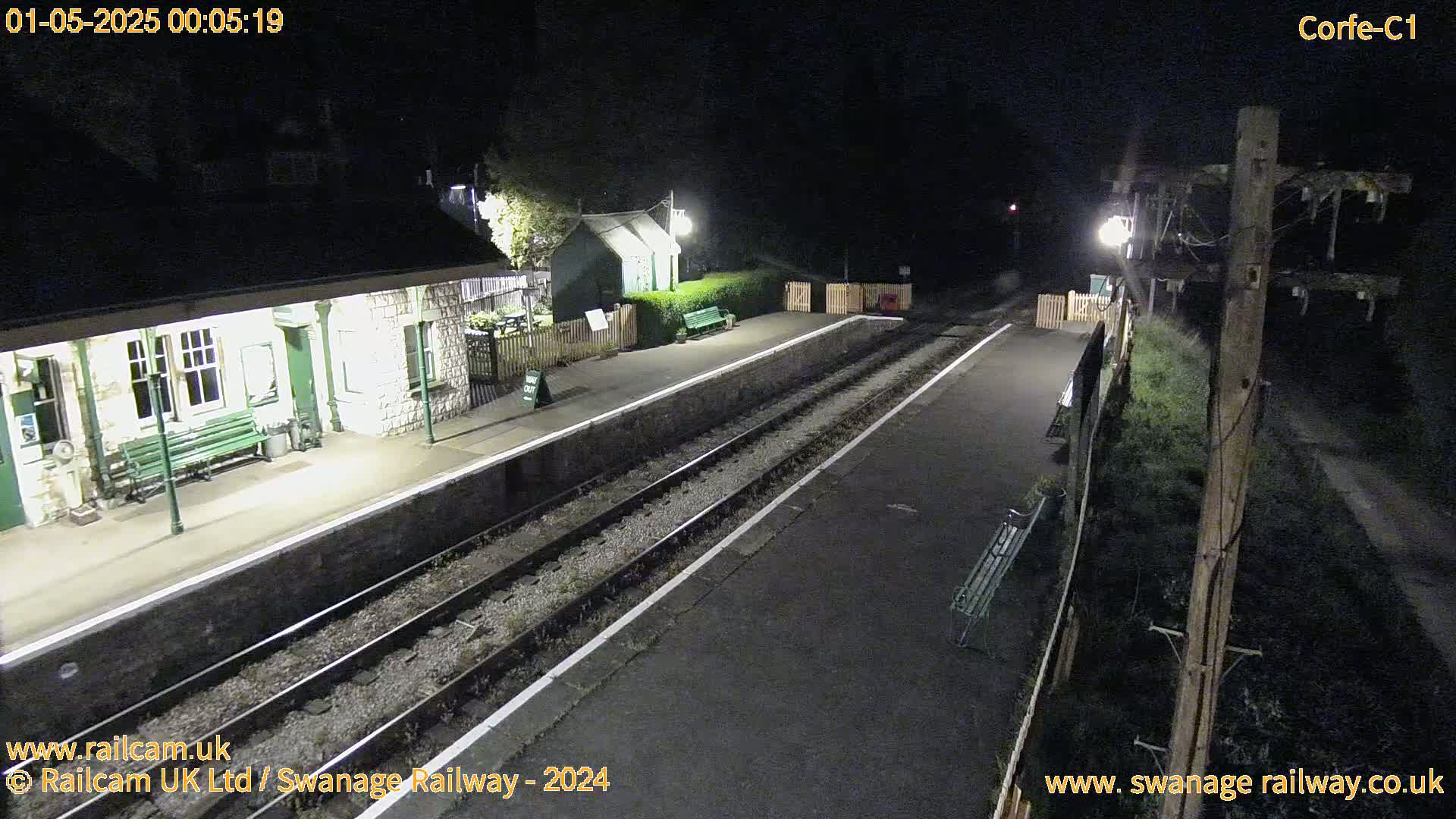 A nighttime view of a deserted train station platform with two sets of tracks, lit by artificial lights.