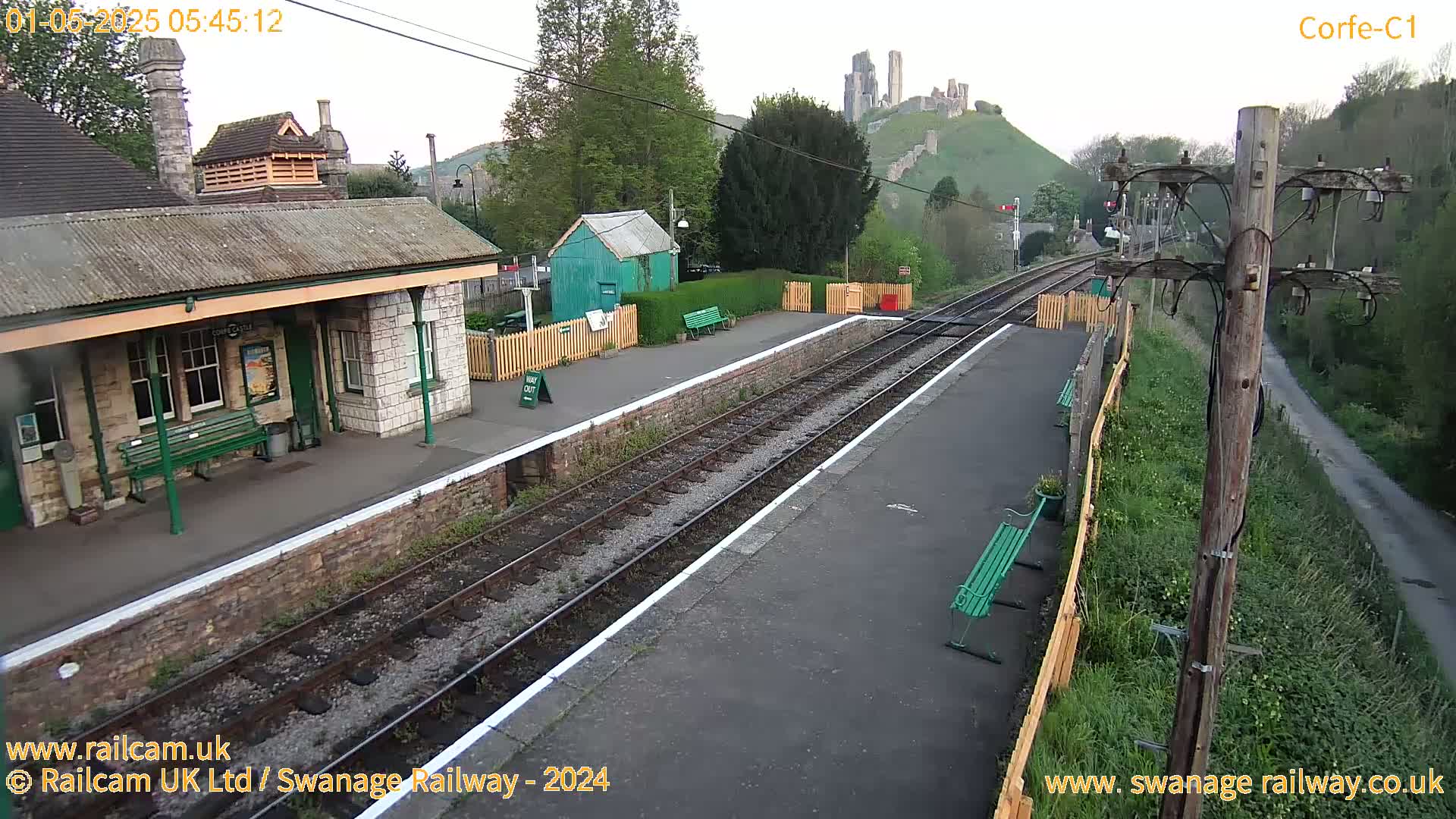 A railway station platform with a stone building and benches, overlooking a hill with a castle ruin under an overcast sky.