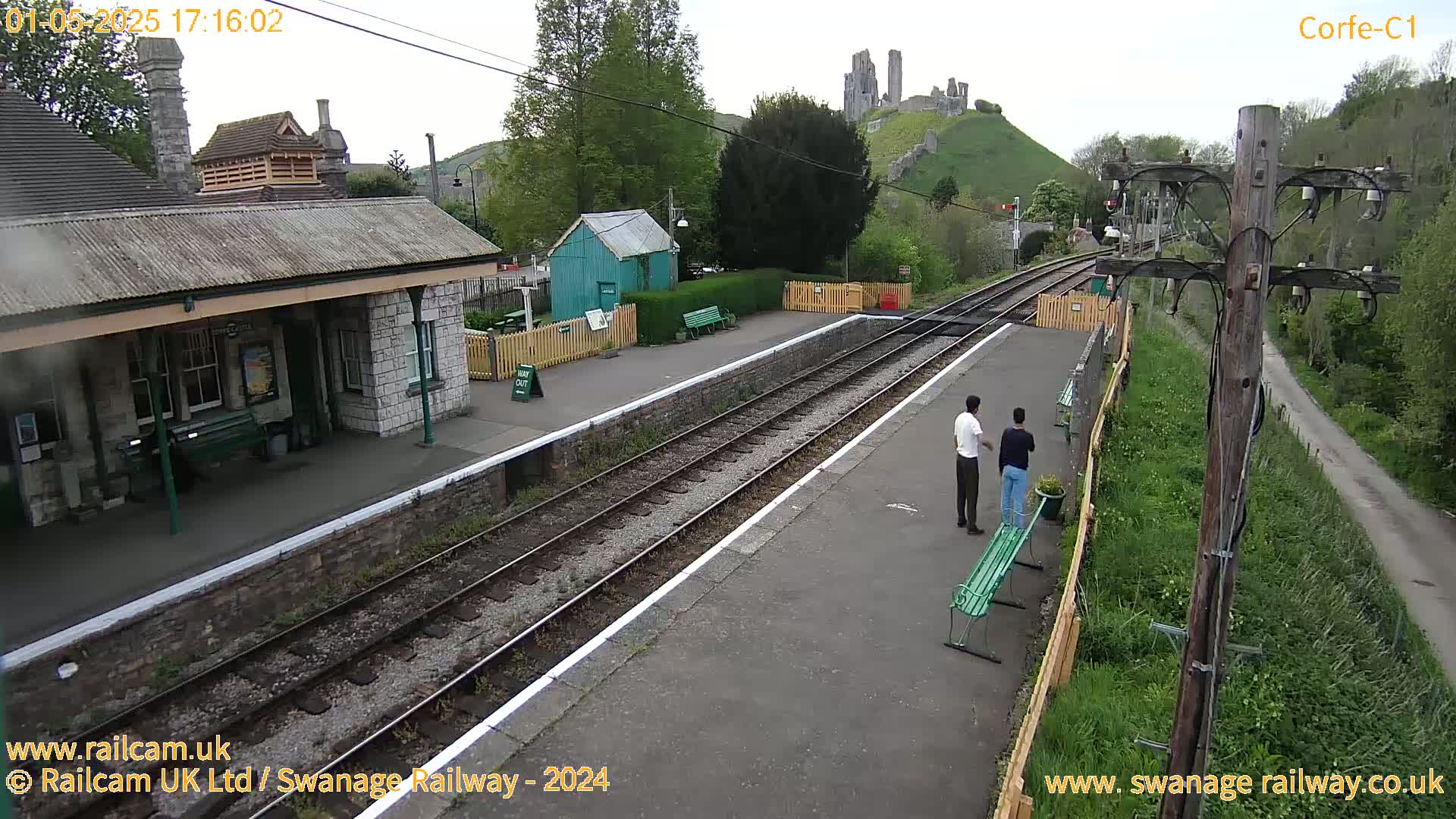 A train station platform with two people standing near a bench, a stone building, and a distant castle visible on a hill under overcast skies.