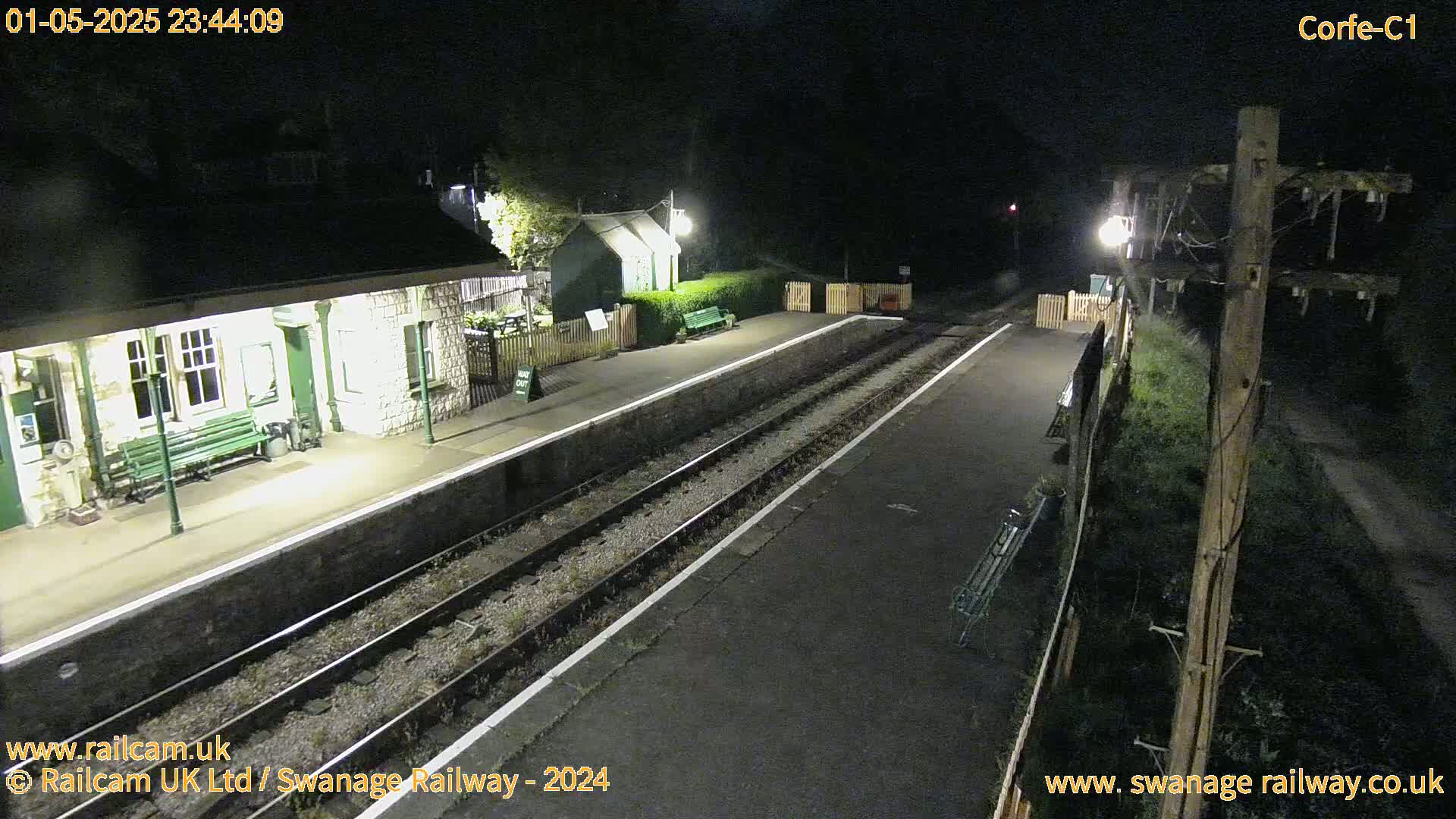 A nighttime view of a deserted train station platform with two sets of train tracks and a stone building illuminated by lights.