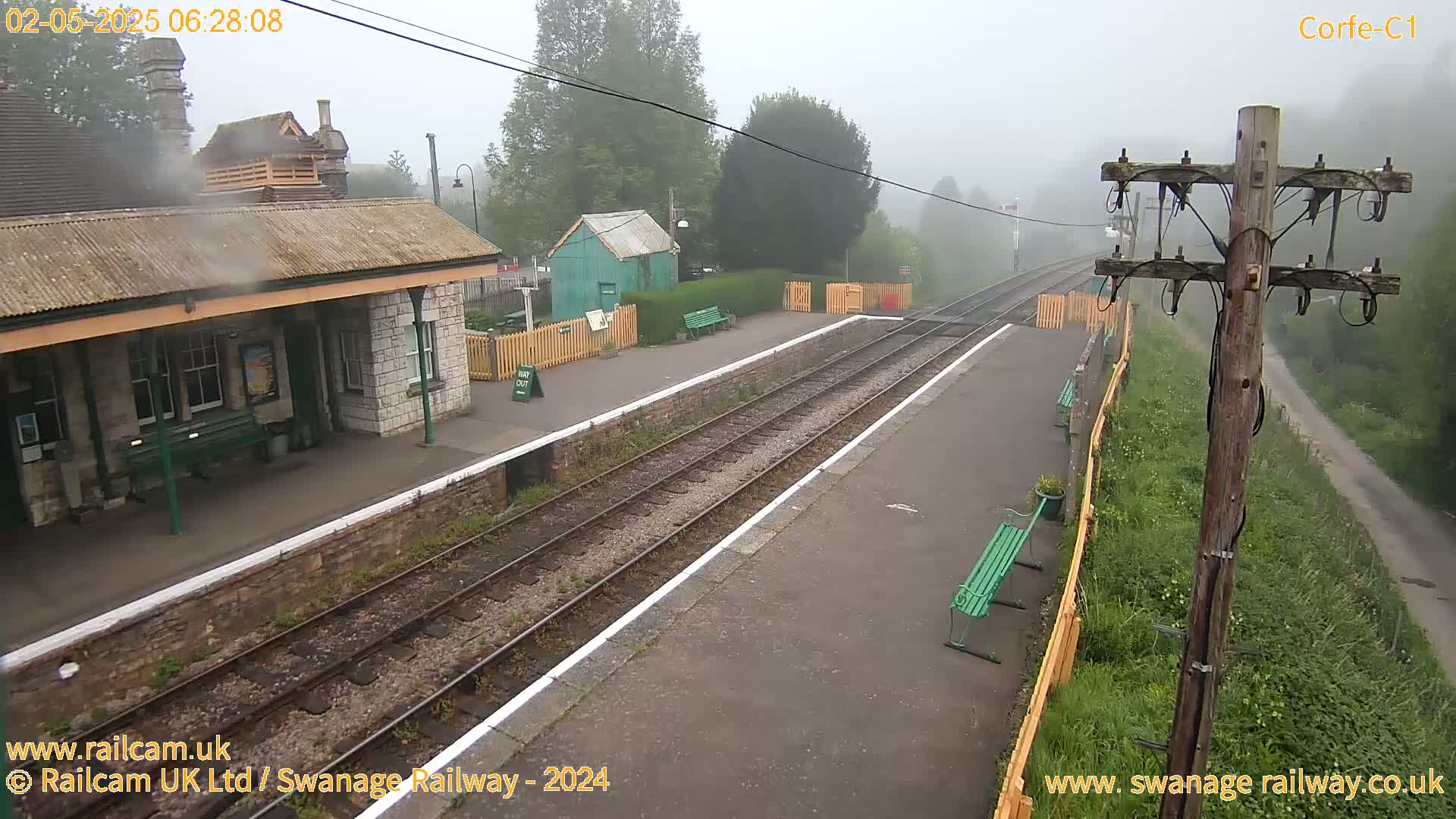 A foggy train station with a stone building, a smaller green building, and several benches sits alongside train tracks.