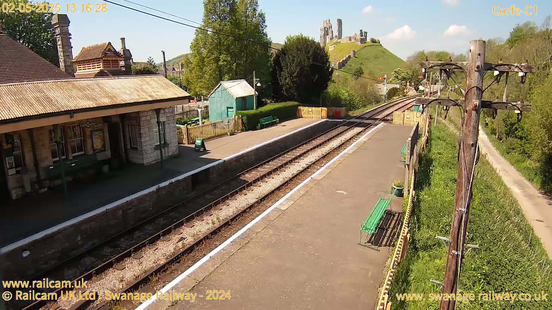 A train station platform with a stone building and a distant castle on a hill, under a sunny sky.