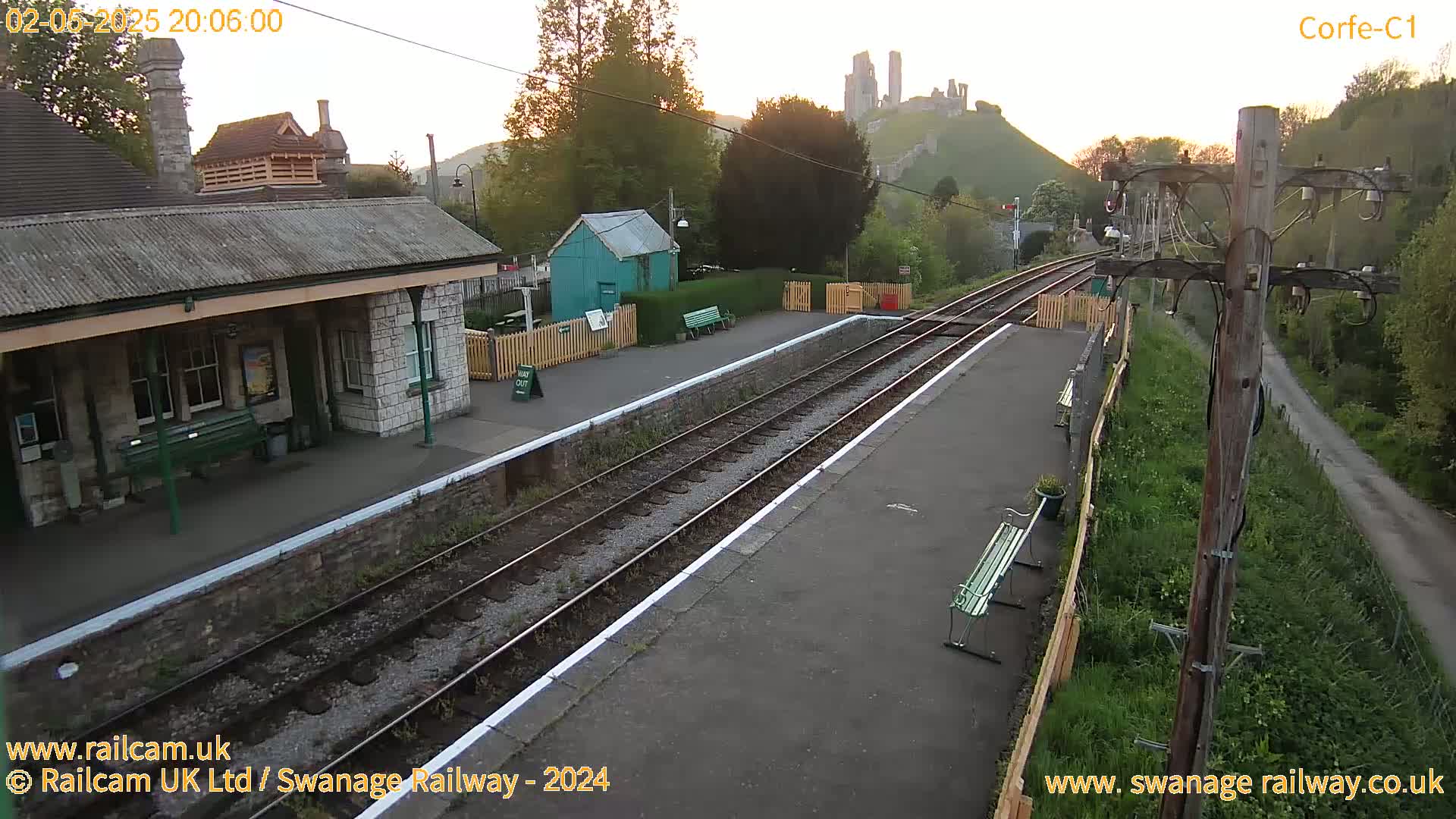 A train station platform at sunrise, with a stone building and a distant castle visible on a hill.