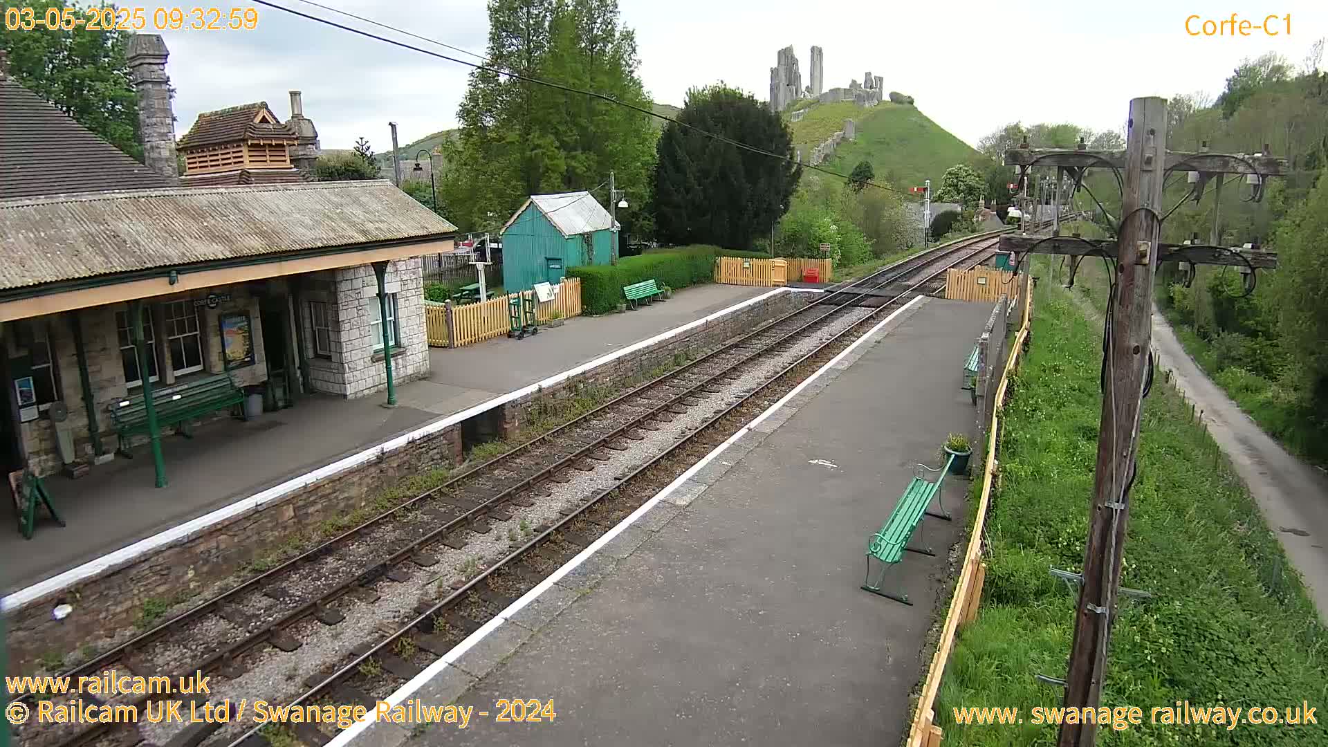 A train station platform with a stone building, a small green shed, and a distant hilltop castle is visible under an overcast sky.