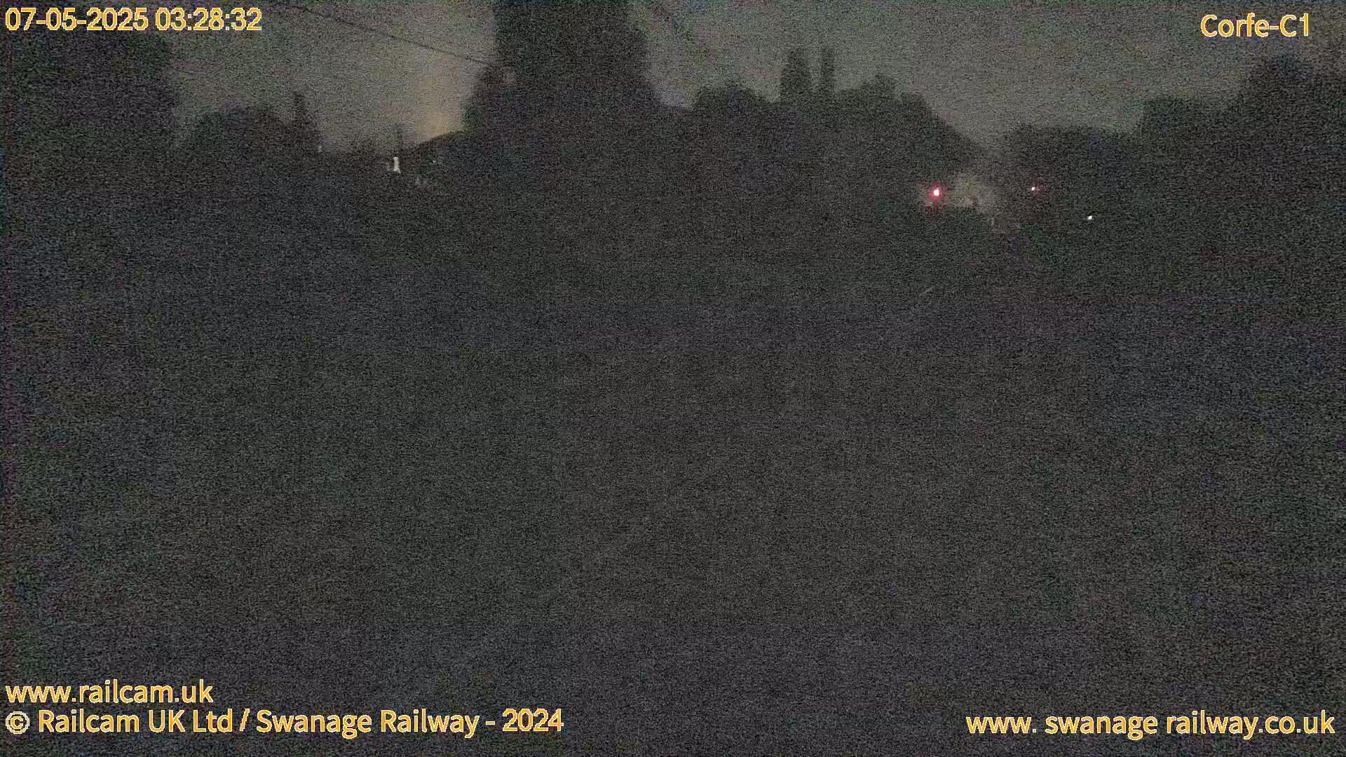A nighttime, low-light outdoor view shows dark vegetation and indistinct structures in the distance under a cloudy sky.