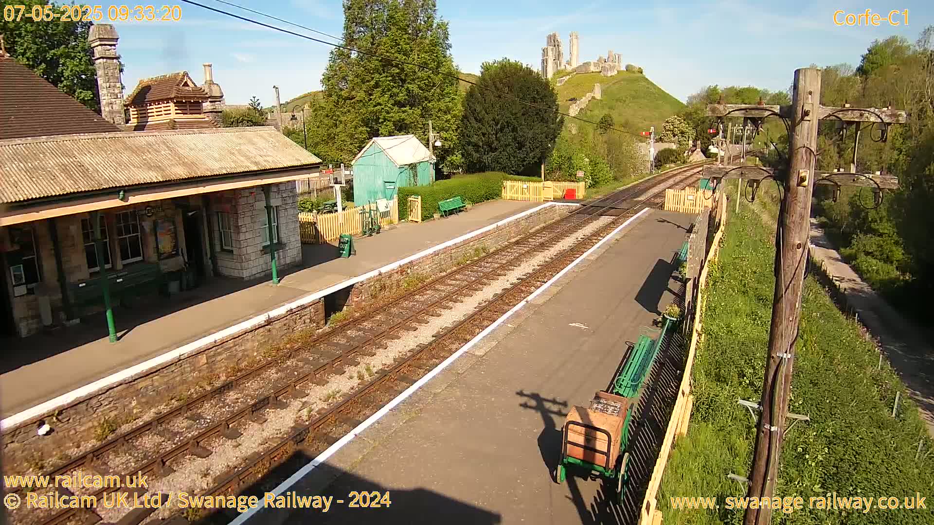 A train station platform on a sunny day with a stone building, a small green shed, and a hilltop castle in the background.
