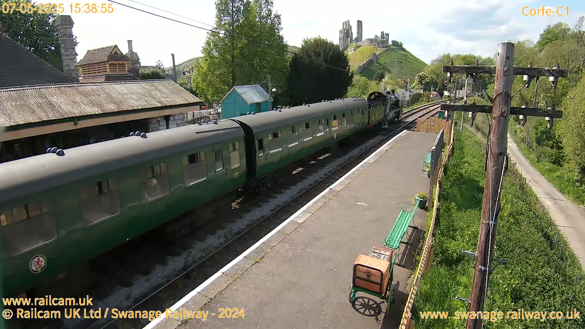 A green passenger train pulls into a railway station on a sunny day, with a castle visible in the distance.