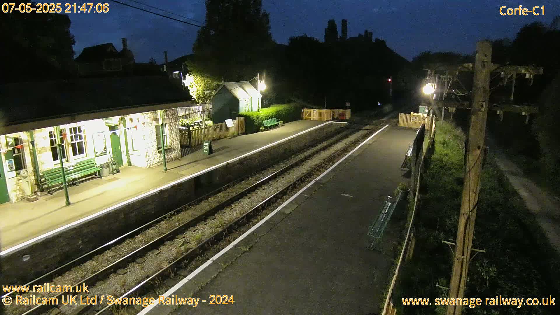 A deserted, nighttime train platform with two sets of tracks, a stone station building, and surrounding vegetation under a dark sky.