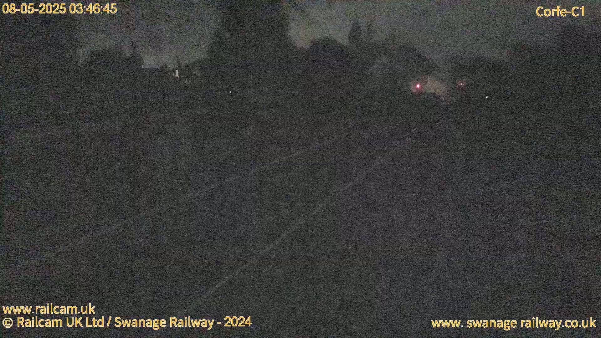 A dark, nighttime view of a railway track, with faint lights visible in the distance under a cloudy sky.