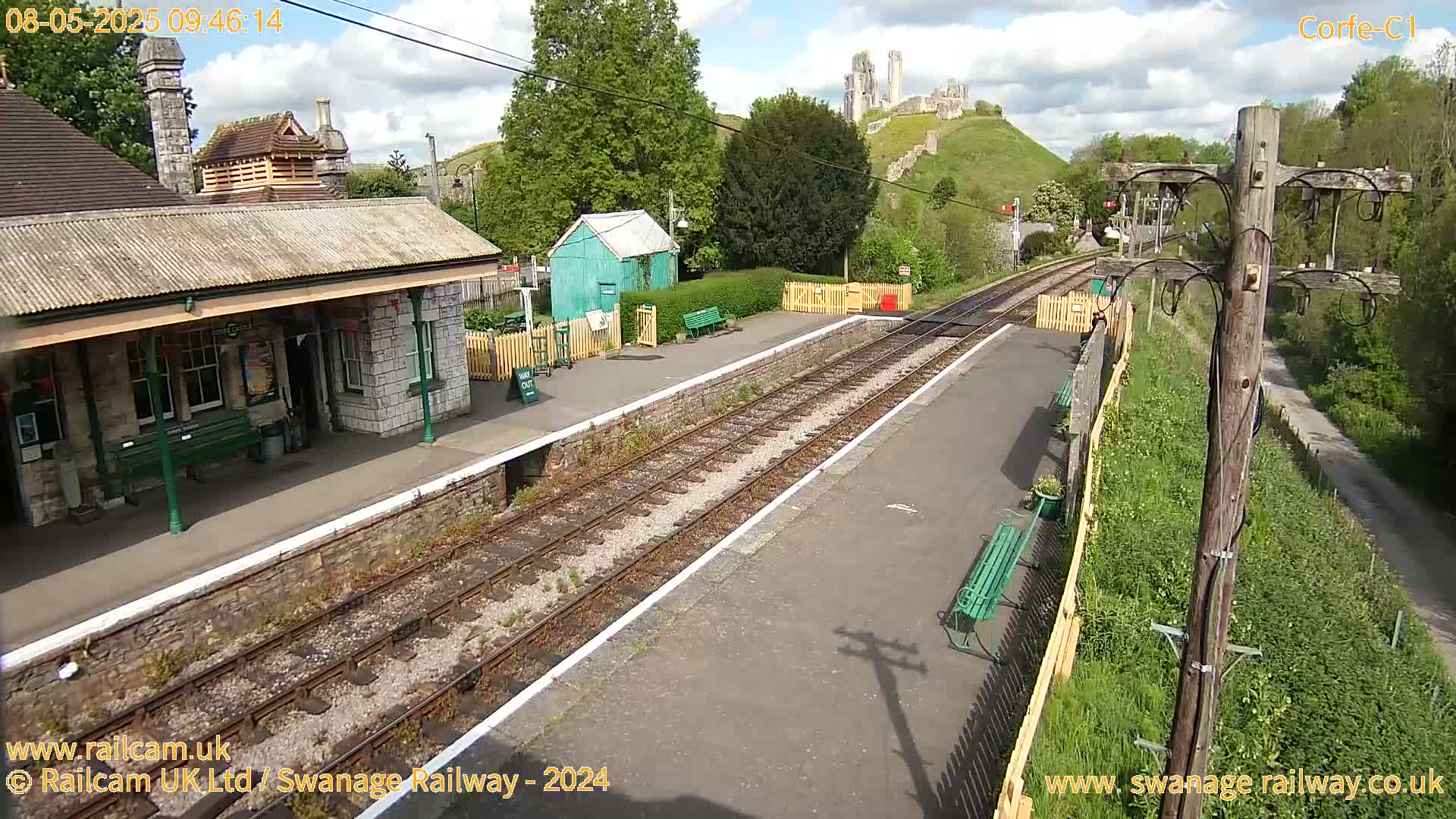 A train station platform with a stone building, a small green shed, and railway tracks stretches towards a hilltop with castle ruins under a partly cloudy sky.