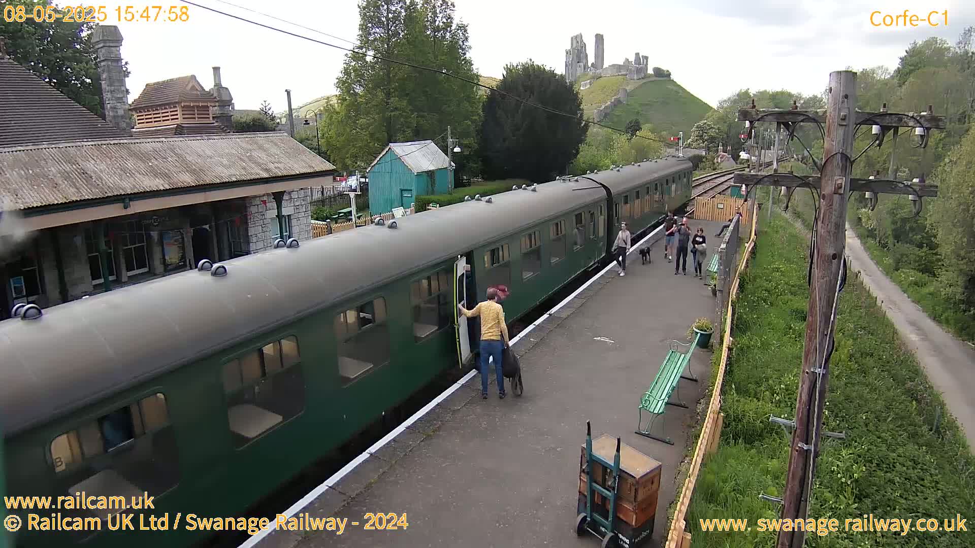 A green passenger train is at a station platform with several people disembarking, under a partly cloudy sky, with a stone ruin visible on a hill in the background.