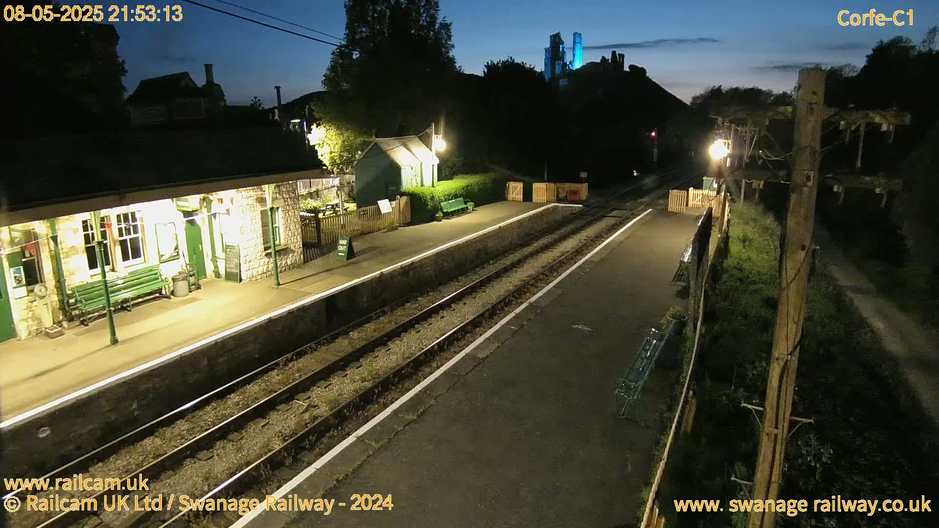 A nighttime view of a train station platform with a stone building, a small green shed, and a castle visible in the distance under a twilight sky.