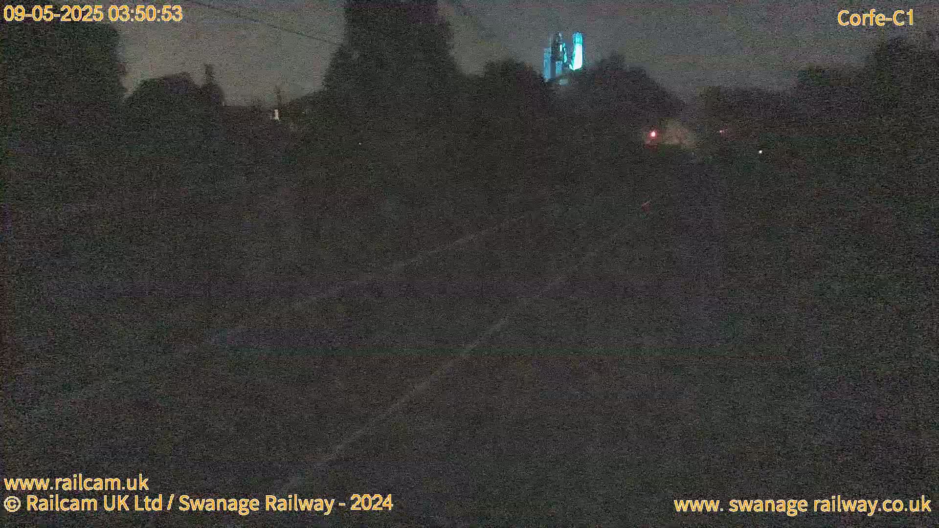 A dark, nighttime view of a railway track, with a faintly illuminated structure visible in the distance.