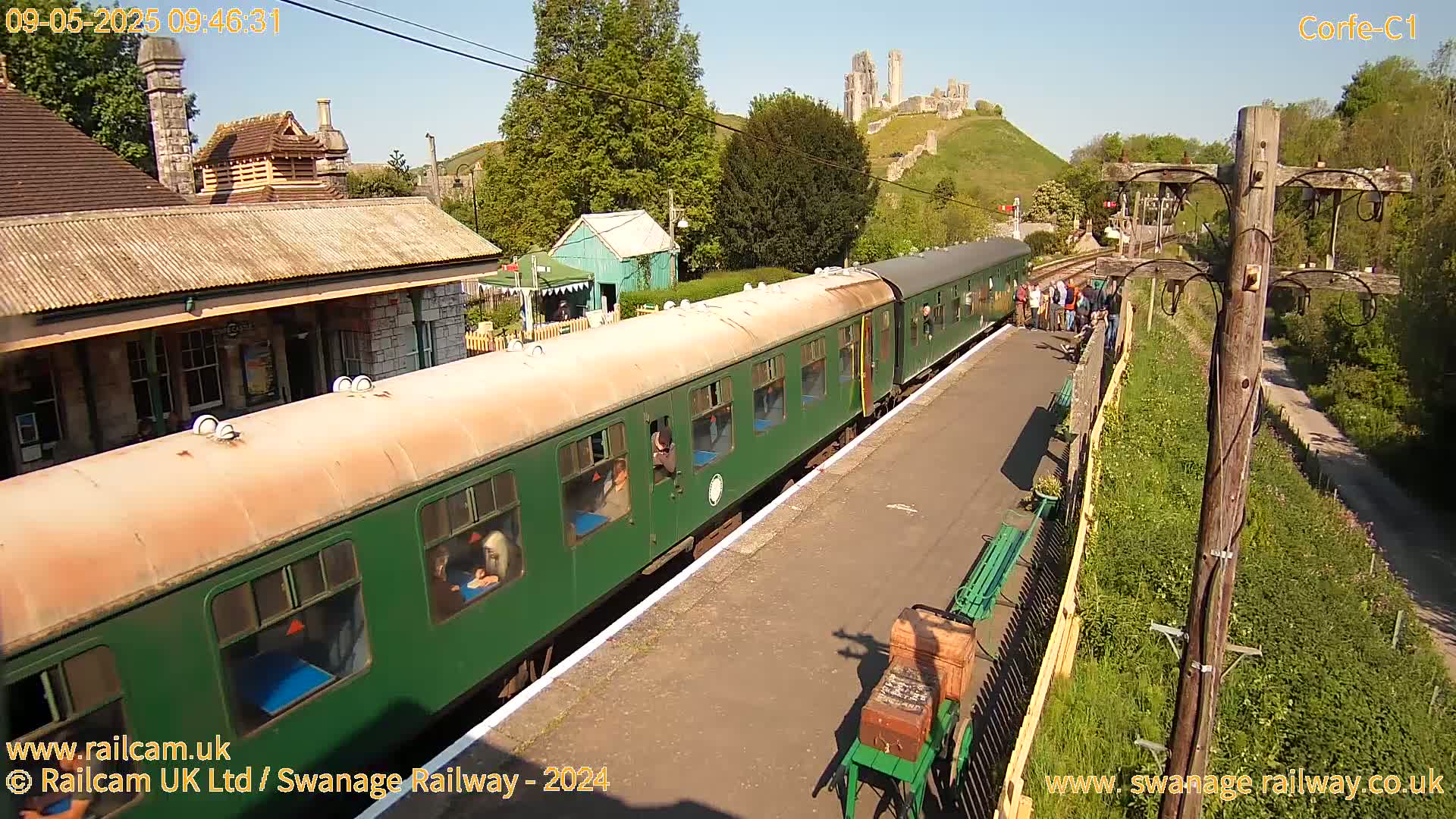 A green passenger train is at a station platform on a sunny day, with a stone castle visible on a hill in the background.