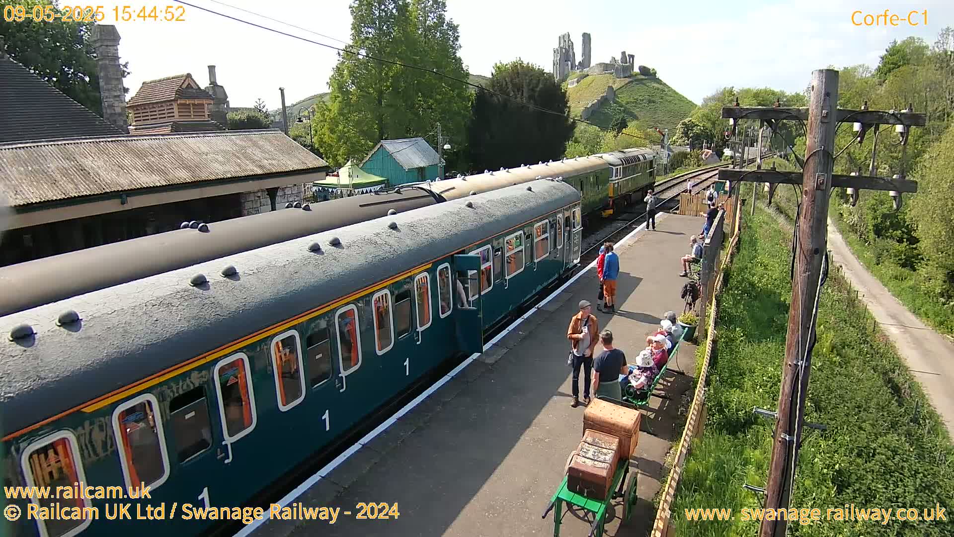 A teal passenger train is stopped at a station platform with people waiting and a castle ruin visible on a hill in the background on a sunny day.
