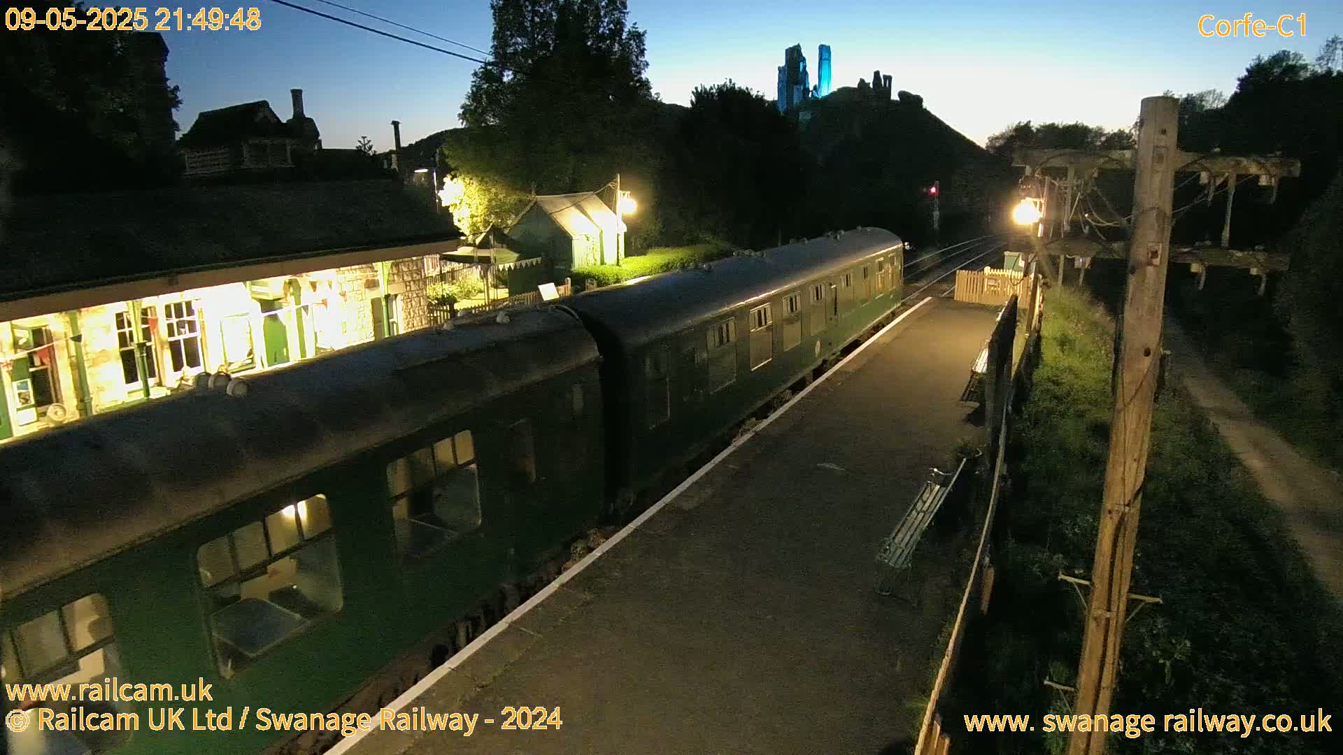 A dark green passenger train sits at a brightly lit station at twilight, with a castle visible in the distance.