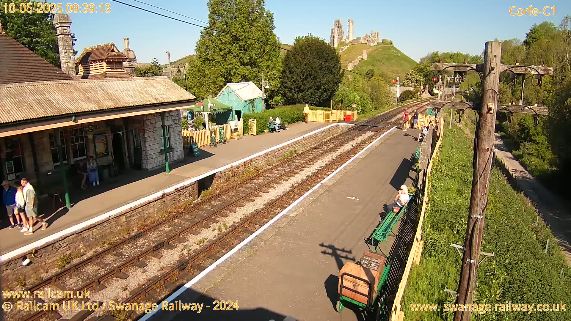A train station platform with a stone building and a castle ruin visible in the distance on a sunny day.
