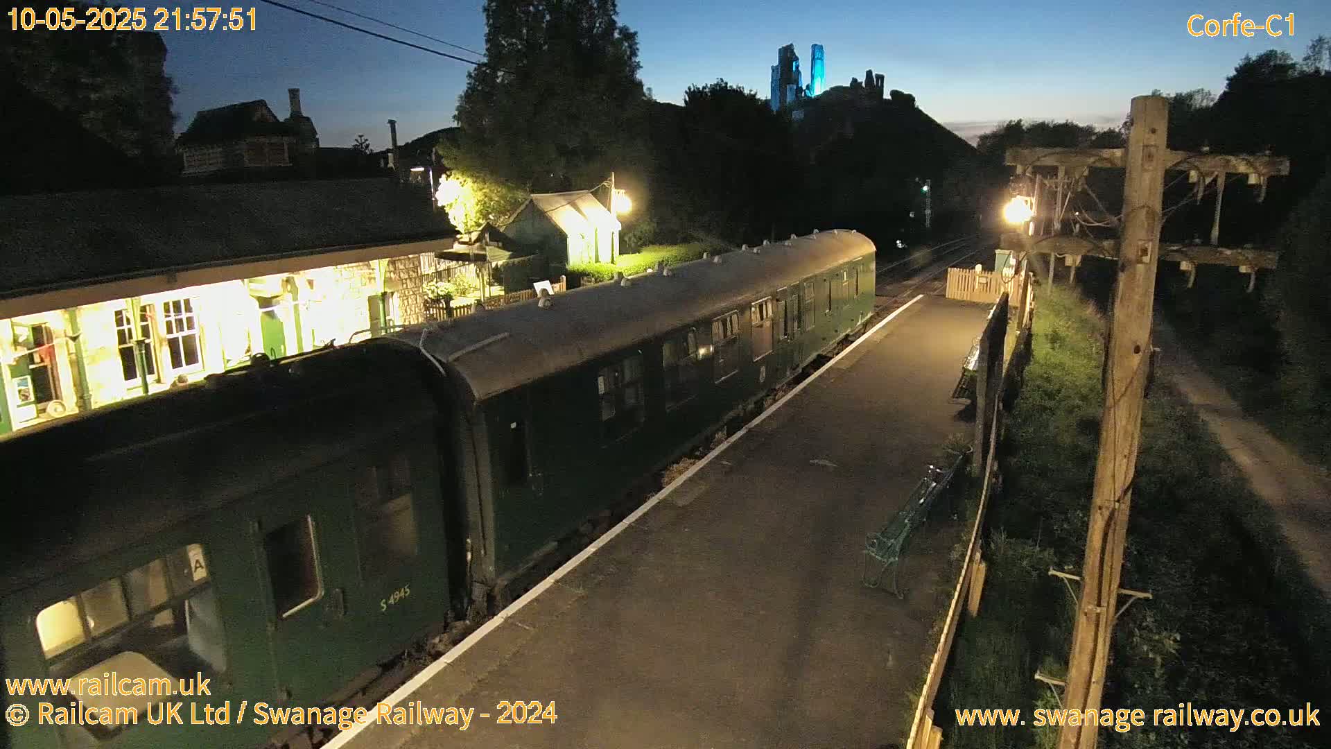 A dark-green passenger train is at a dimly lit train station at night, with a castle visible in the distance under a twilight sky.