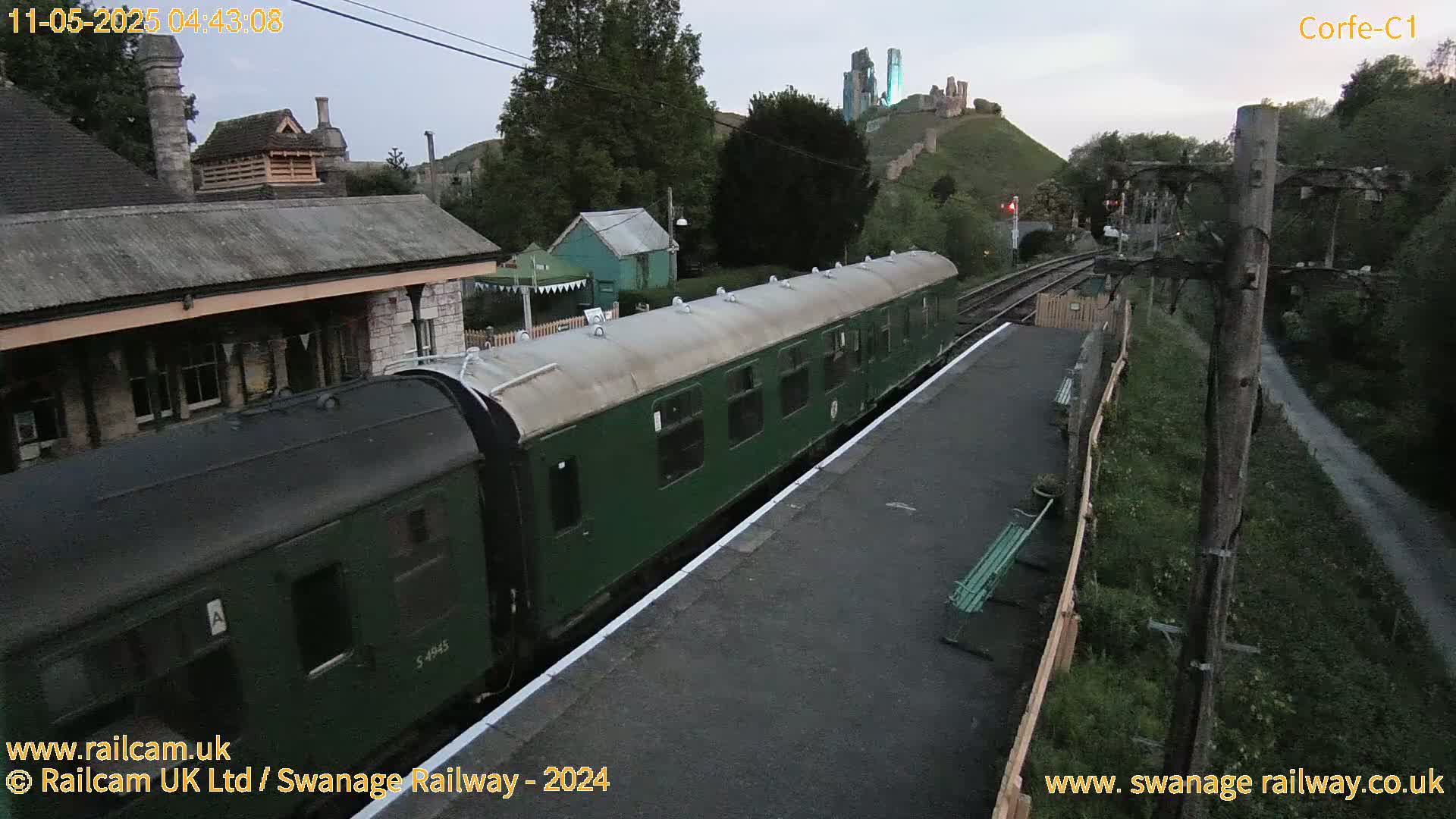 A dark green passenger train is at a station, with a castle visible on a hill in the background under a twilight sky.