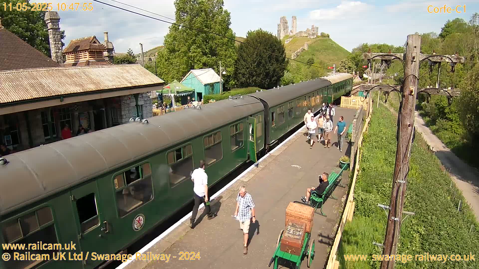 A green passenger train is at a station on a sunny day, with a medieval castle visible in the background.