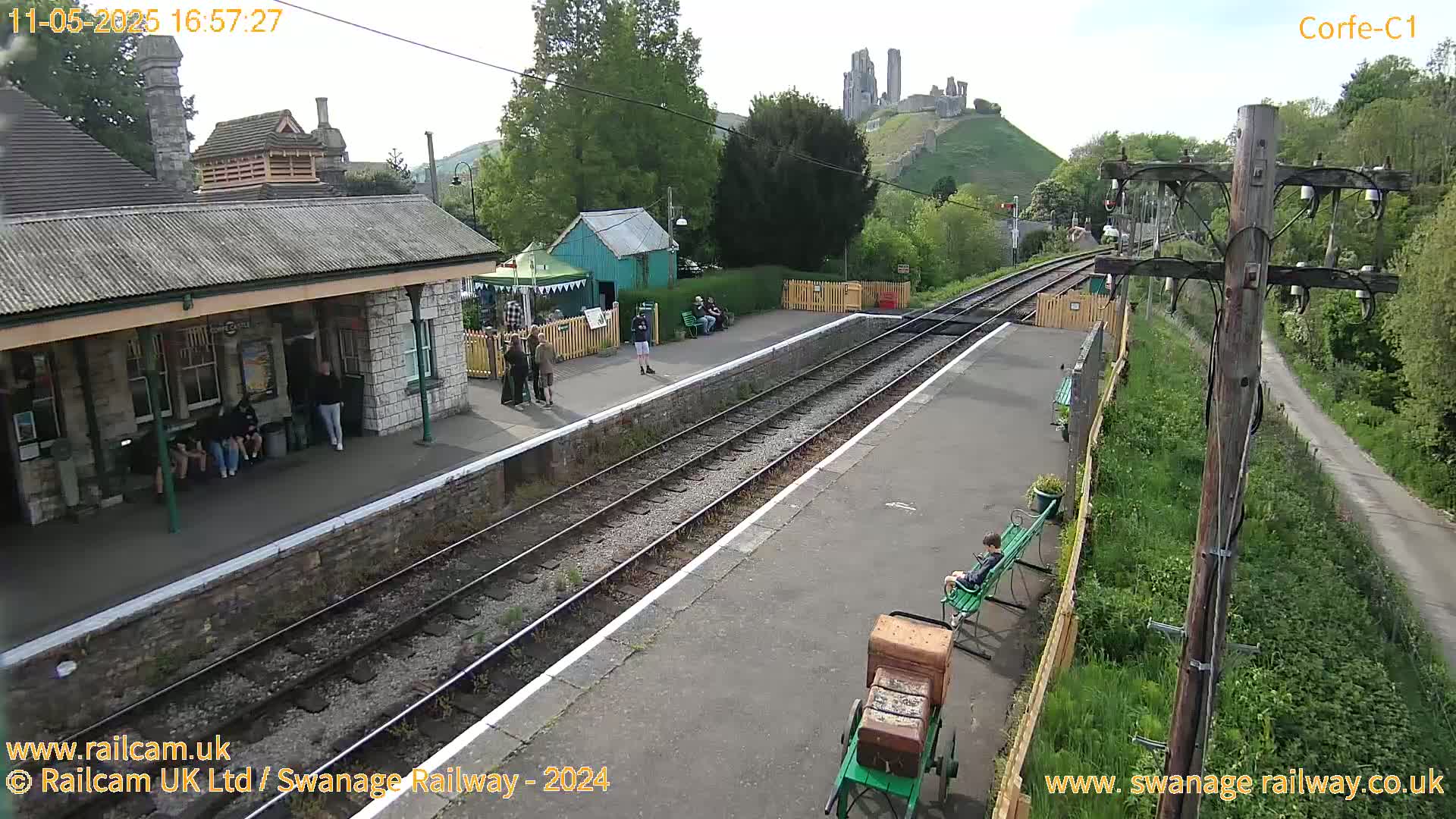 A train station platform with several people waiting, a luggage cart, and a distant hilltop castle is visible under a partly cloudy sky.