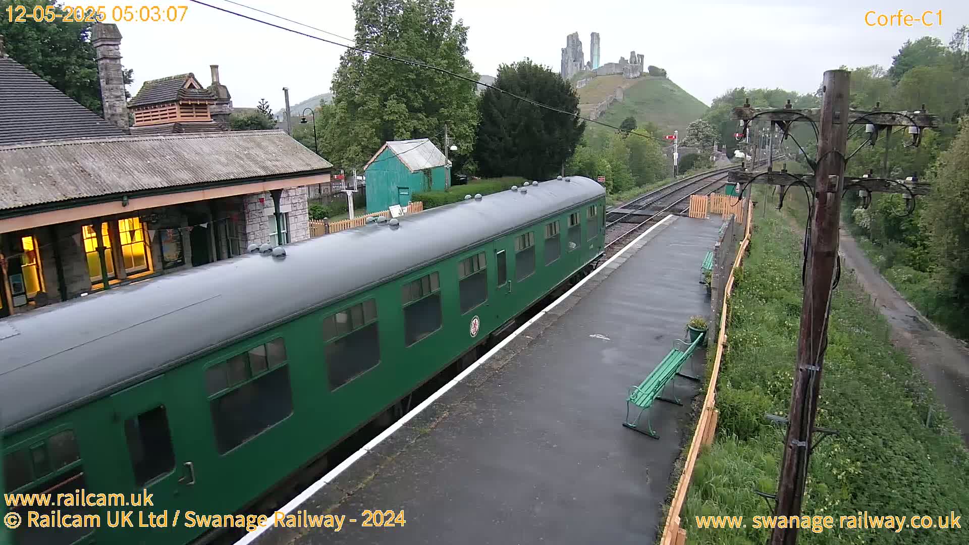 A green passenger train is at a station on a wet, overcast day, with a hilltop castle visible in the background.