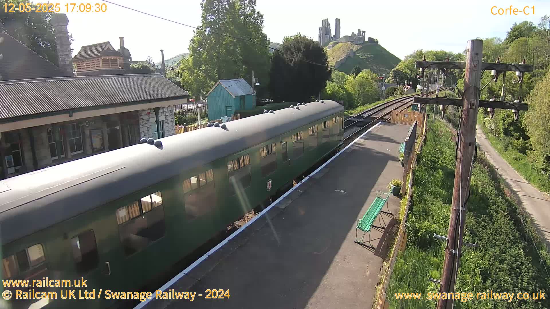 A green passenger train sits at a station on a sunny day, with a castle visible in the distance.