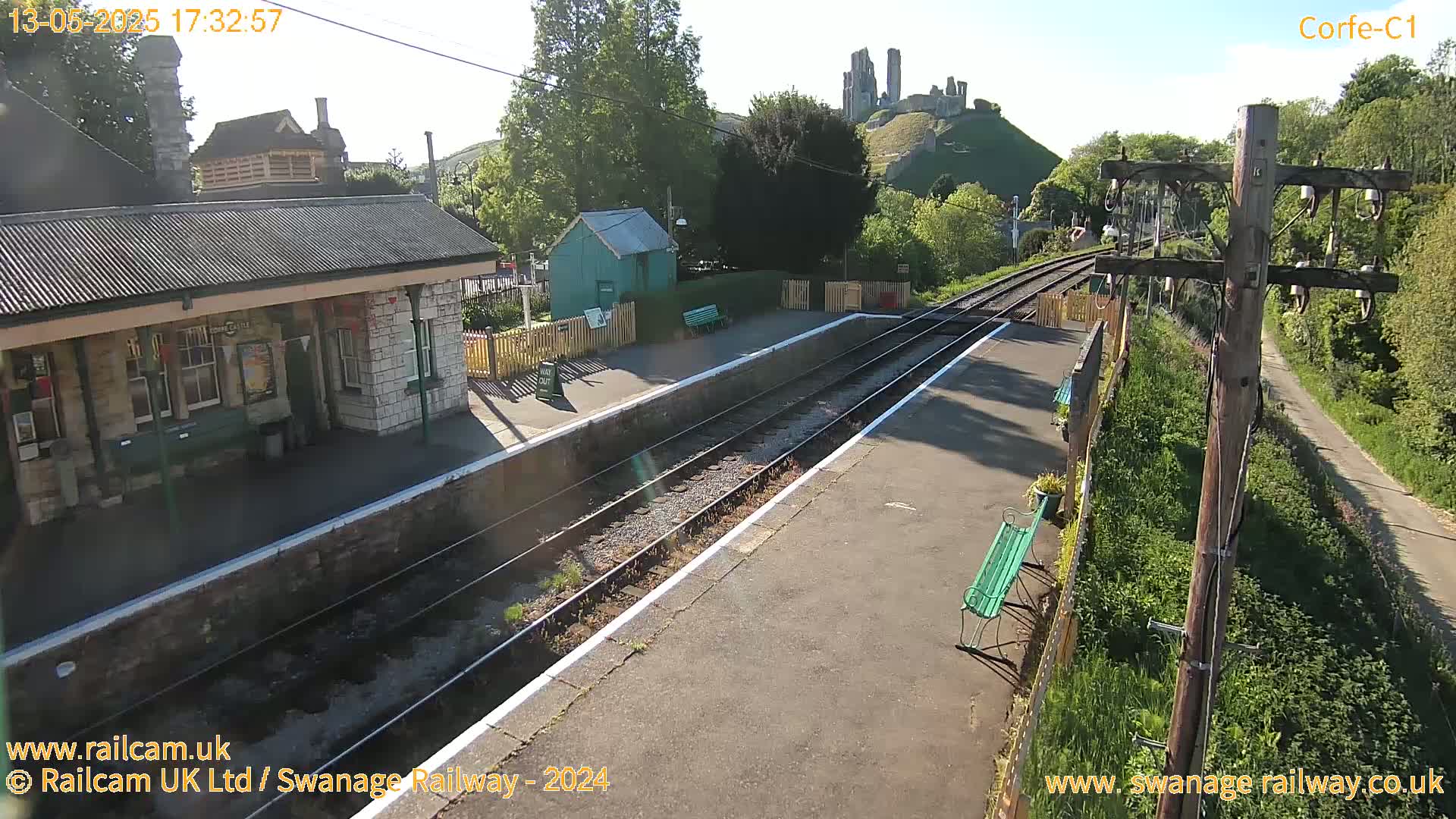 A sunny view of a train station platform with a stone building, a small green structure, and a distant castle visible on a hill.