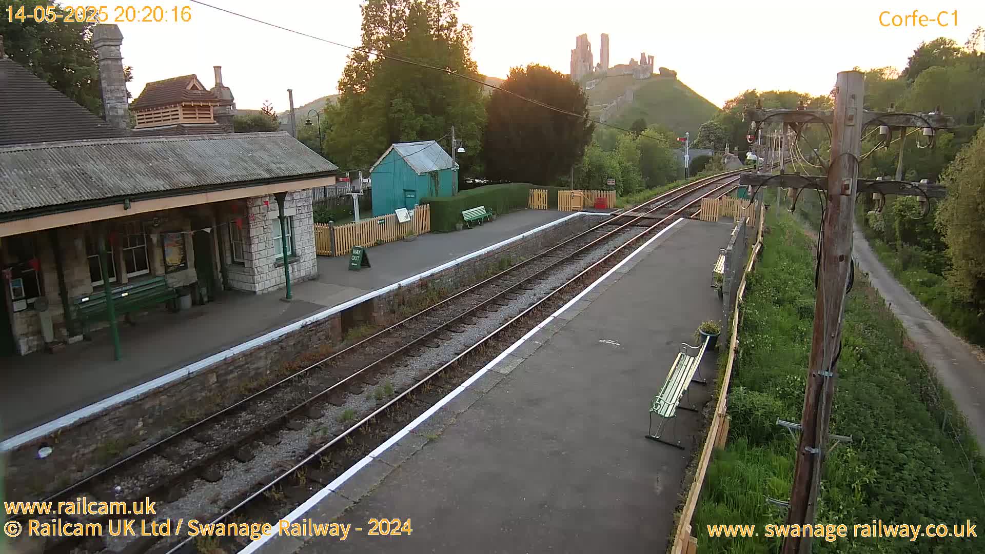 A train station platform with two train tracks, a stone building, and a distant castle is visible at sunset.