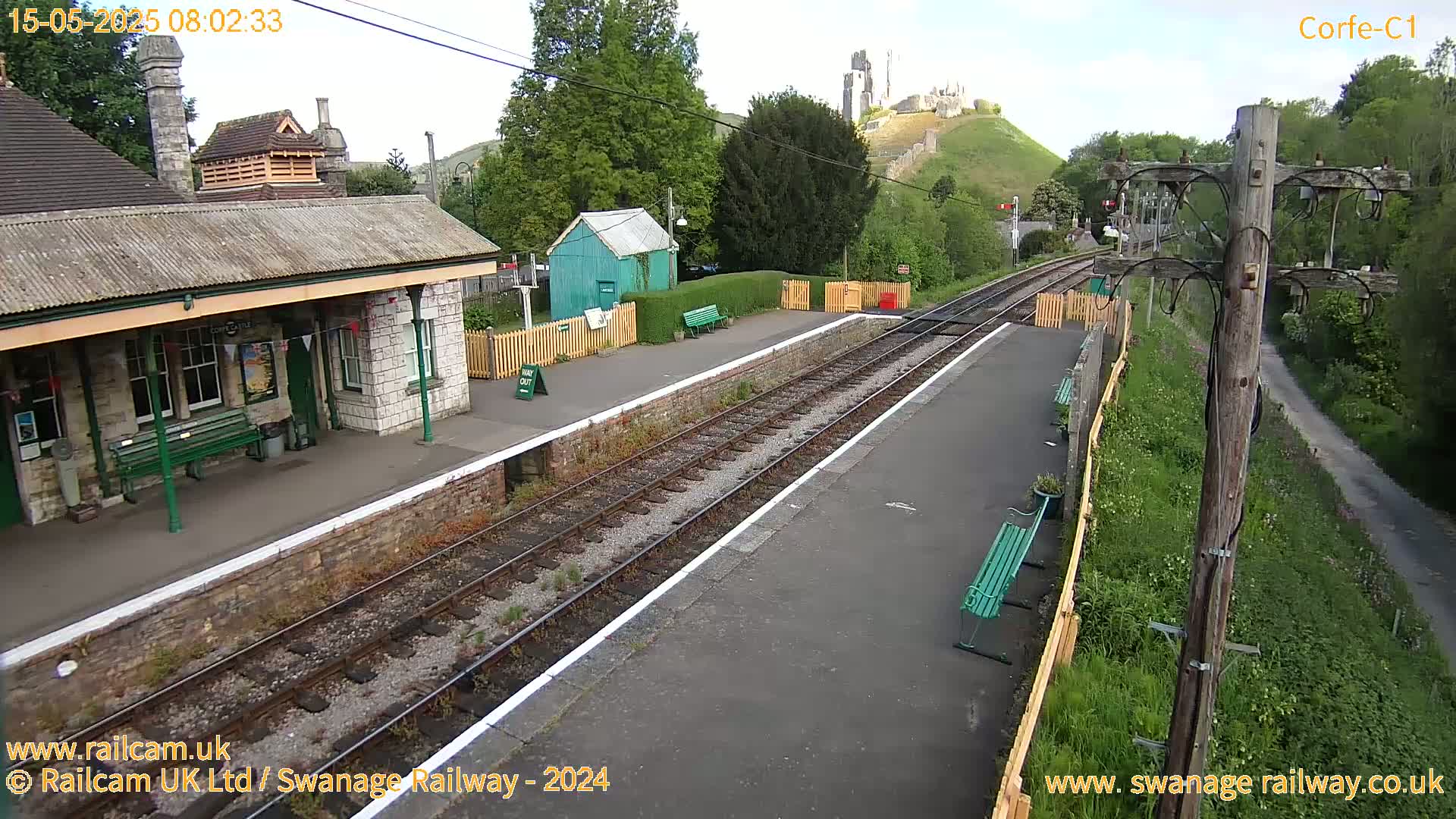 A train station platform with a stone building, a small green shed, and a castle visible on a hill in the background on a partly cloudy day.