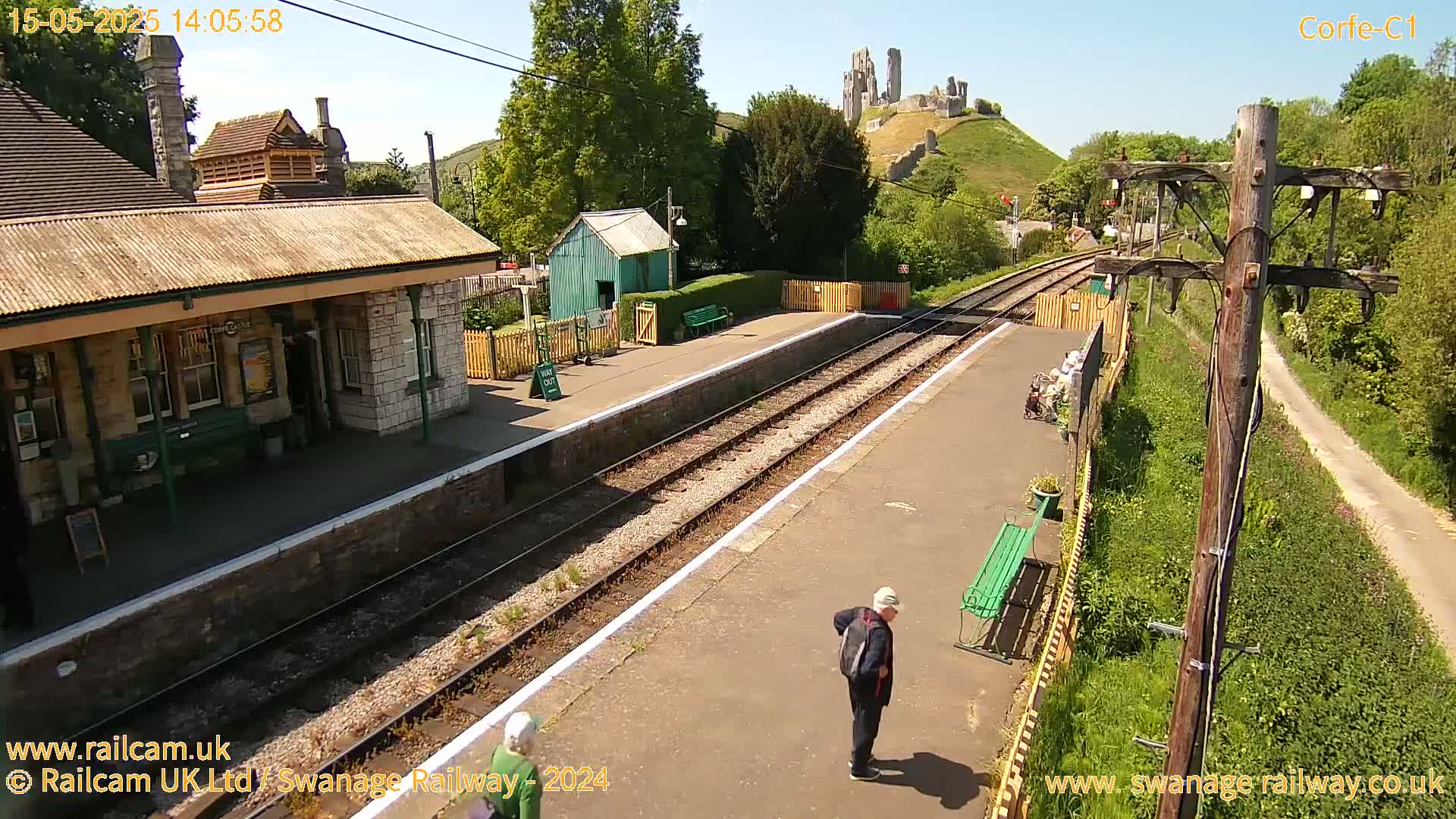 A sunny view of a train station platform with two people standing near a bench, a station building, and a hilltop castle in the background.